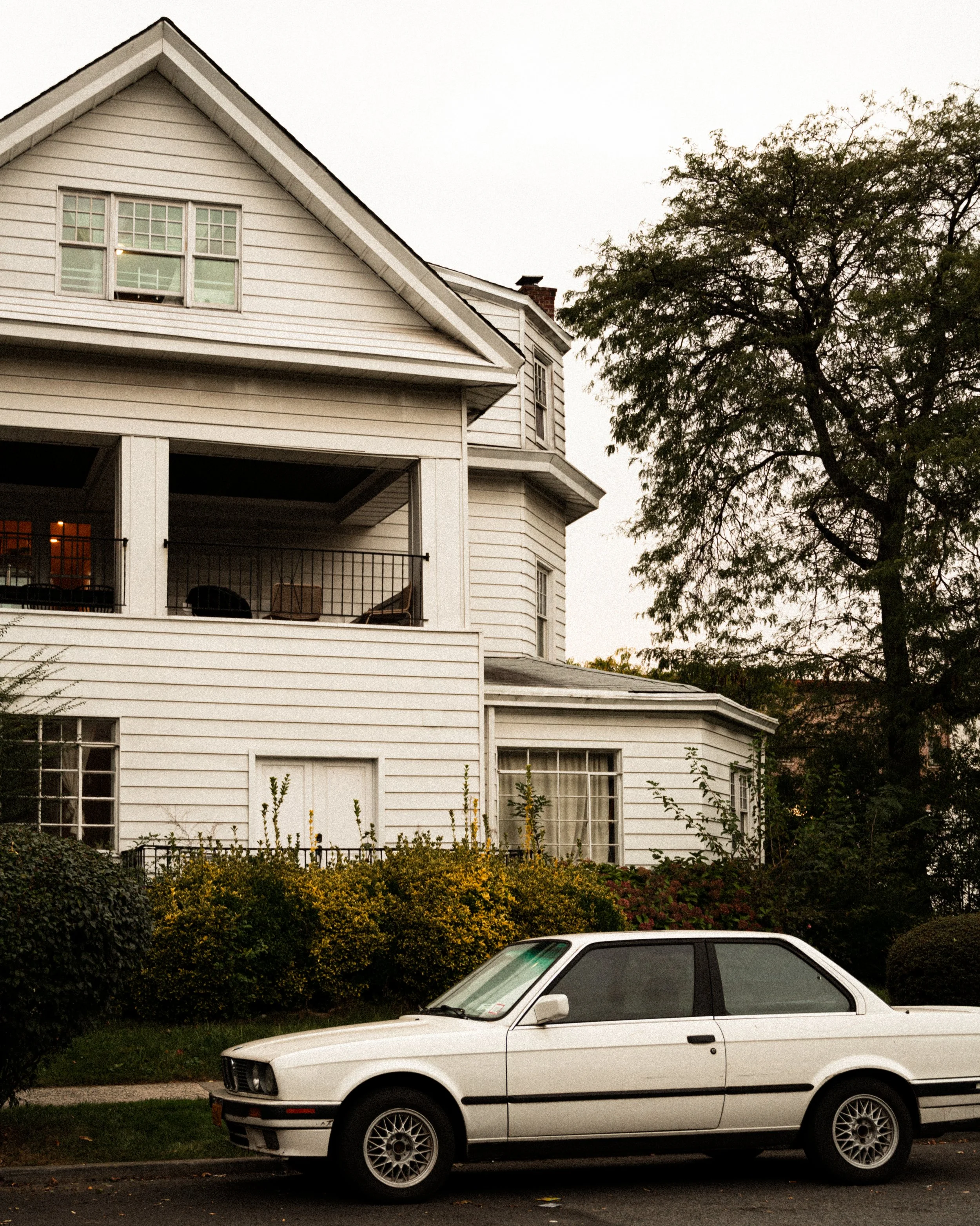 A white vintage car parked in front of a white multi-story house with a porch and surrounding trees and bushes.