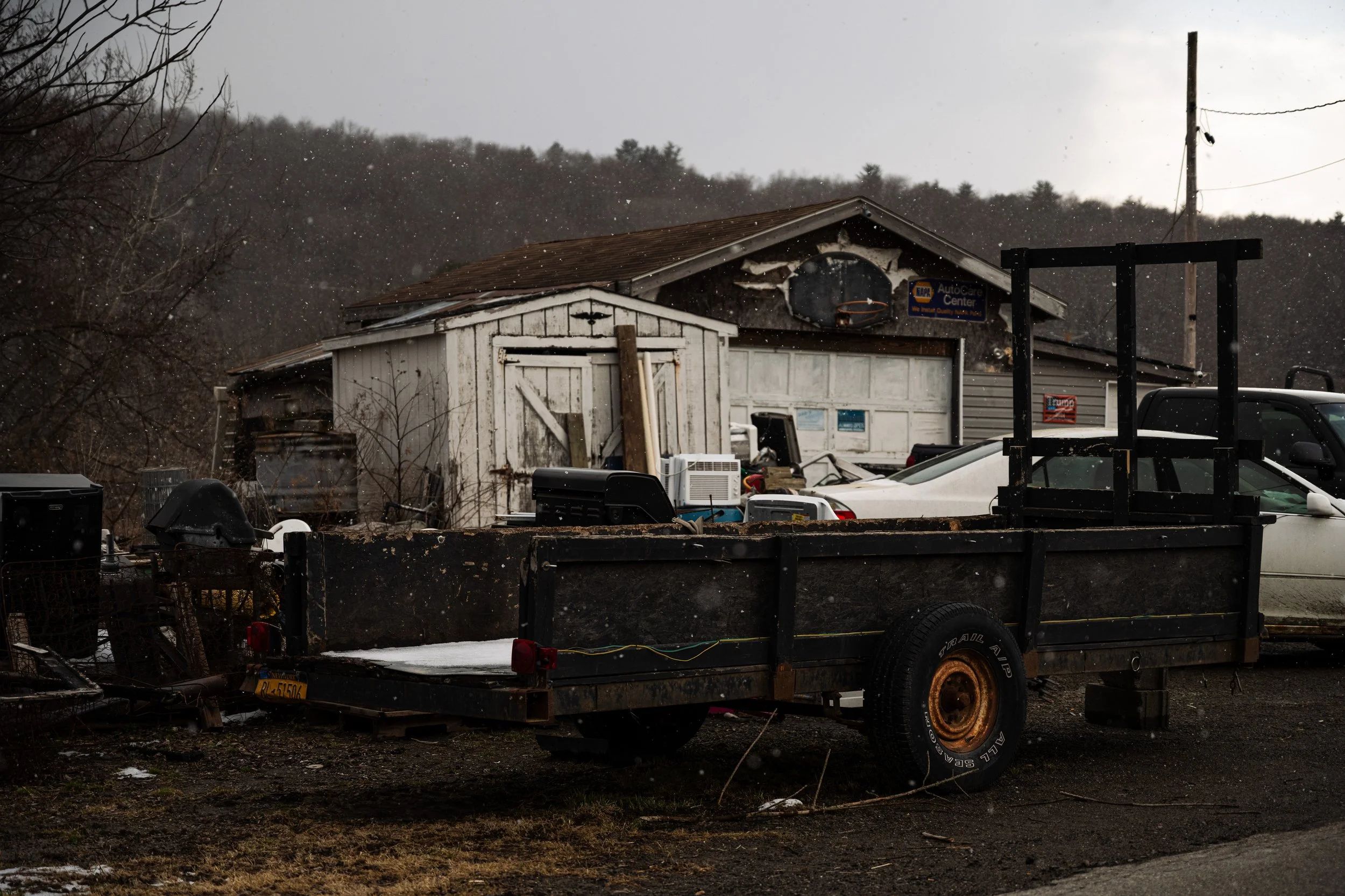 An old, weathered building with a rusted roof and a small shed in front. There is a black flatbed trailer with rusted wheels and various items loaded on it parked nearby. Snow is falling, and the scene appears to be in a rural area.
