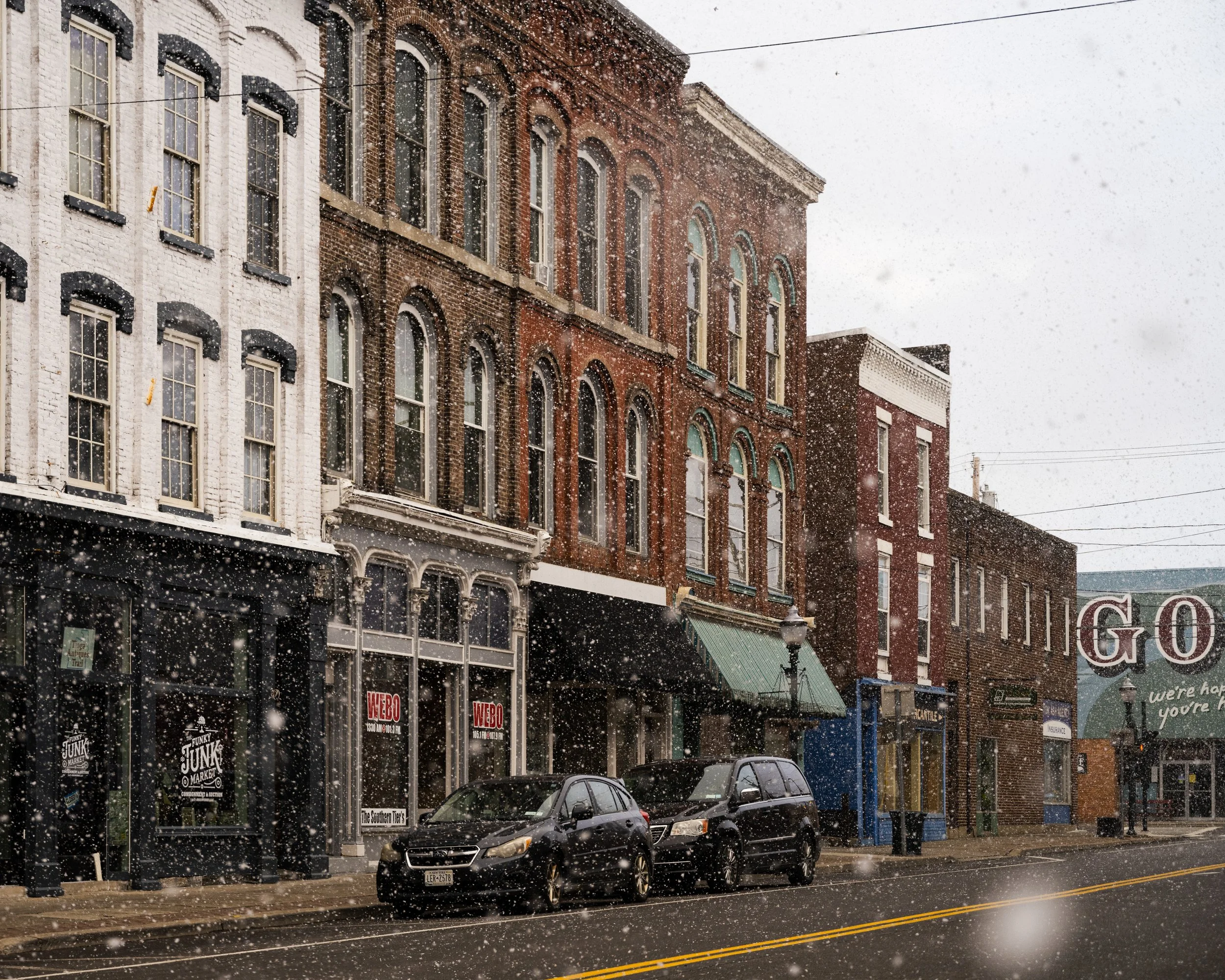 Snow falling on a street lined with historic brick buildings, some with storefronts and parked cars in the foreground. Owego, NY. Upstate NY. Documentary travel art photographer.