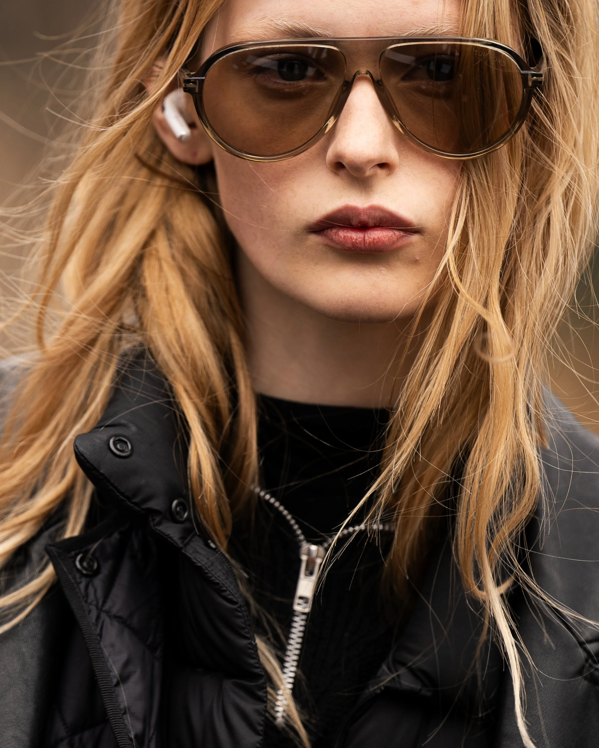 Close-up of a woman with long red hair wearing sunglasses, wireless earbuds, and a black jacket. Street style photograph by street style photographer Kyle Mark Peterson during NYFW.