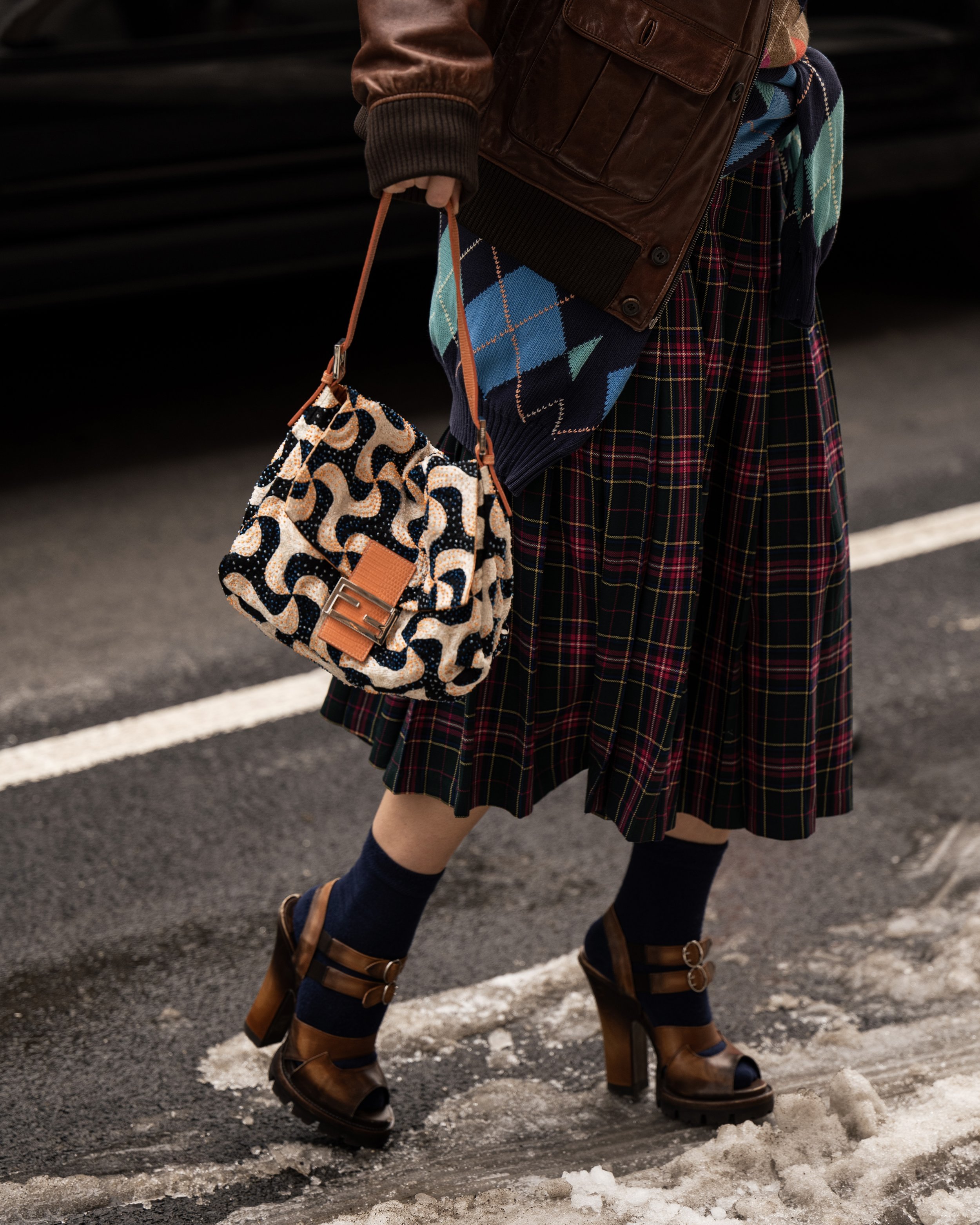 A person standing on a snowy street, wearing a plaid skirt, brown high-heeled sandals, navy socks, and a brown jacket, and carrying a patterned handbag. Street style photograph by street style photographer Kyle Mark Peterson during NYFW.