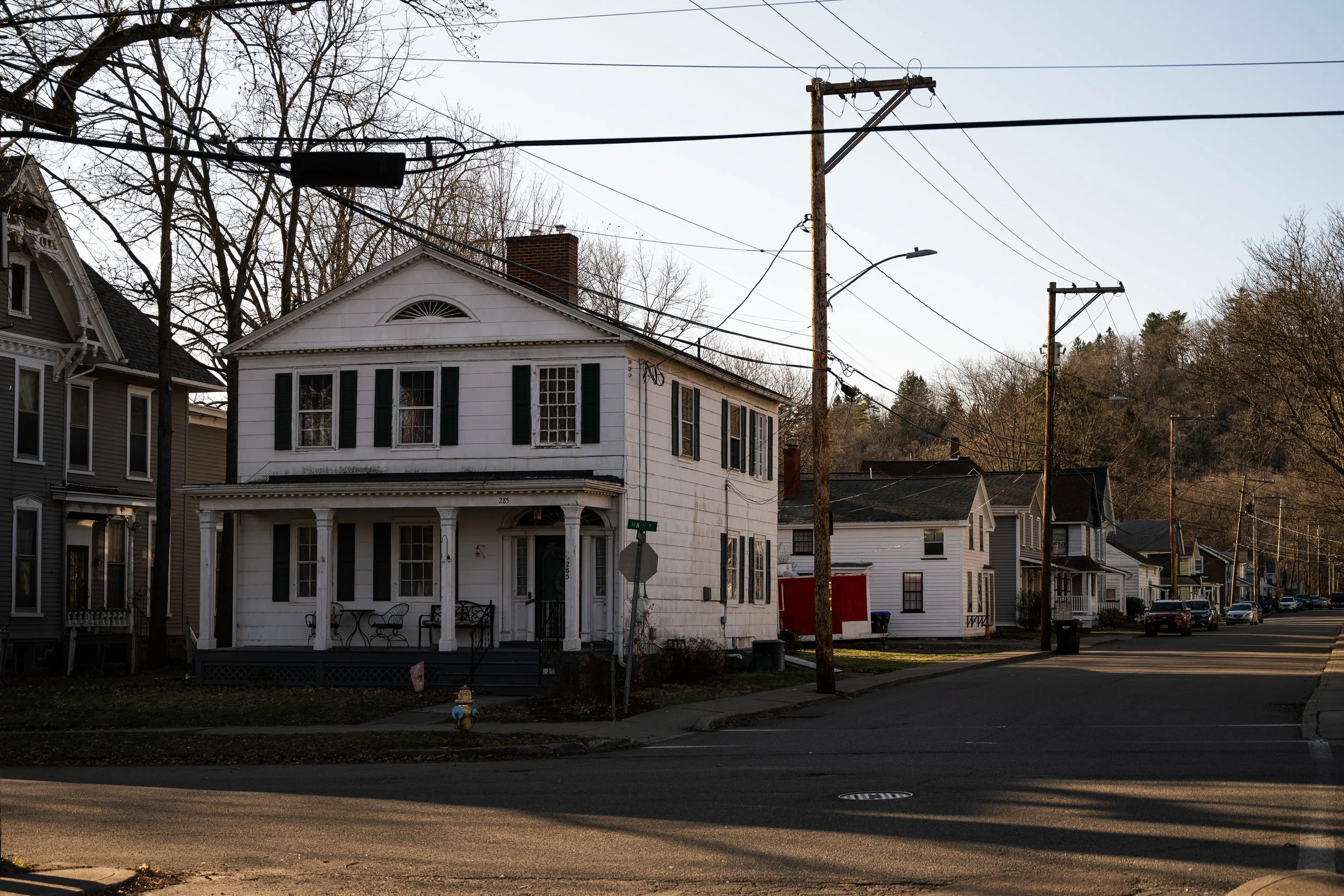 A residential street scene with a two-story white house and neighboring houses, utility poles and power lines, bare trees, and parked cars.