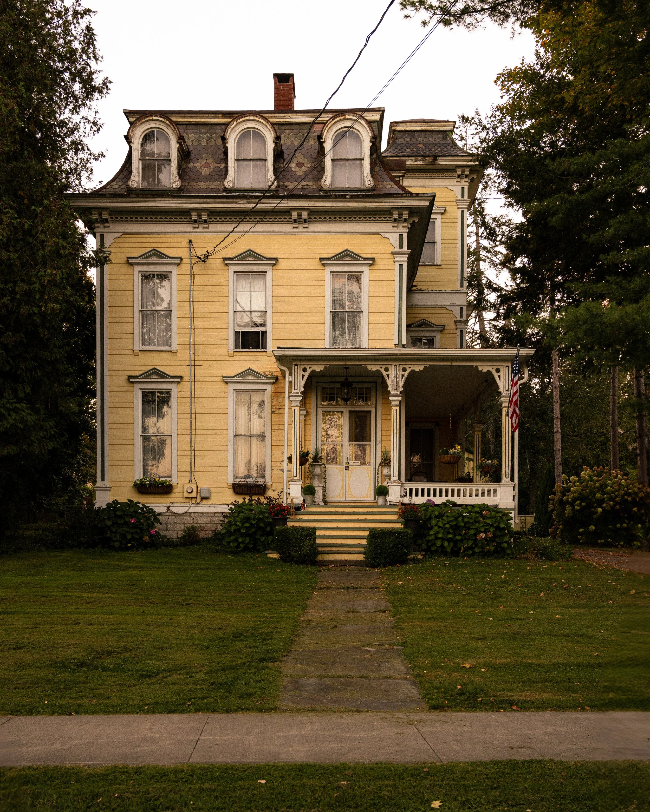 A vintage yellow Victorian house with decorative trim, a porch, and a staircase leading to the front door, surrounded by a well-maintained lawn and trees. Marathon, NY. Upstate New York.