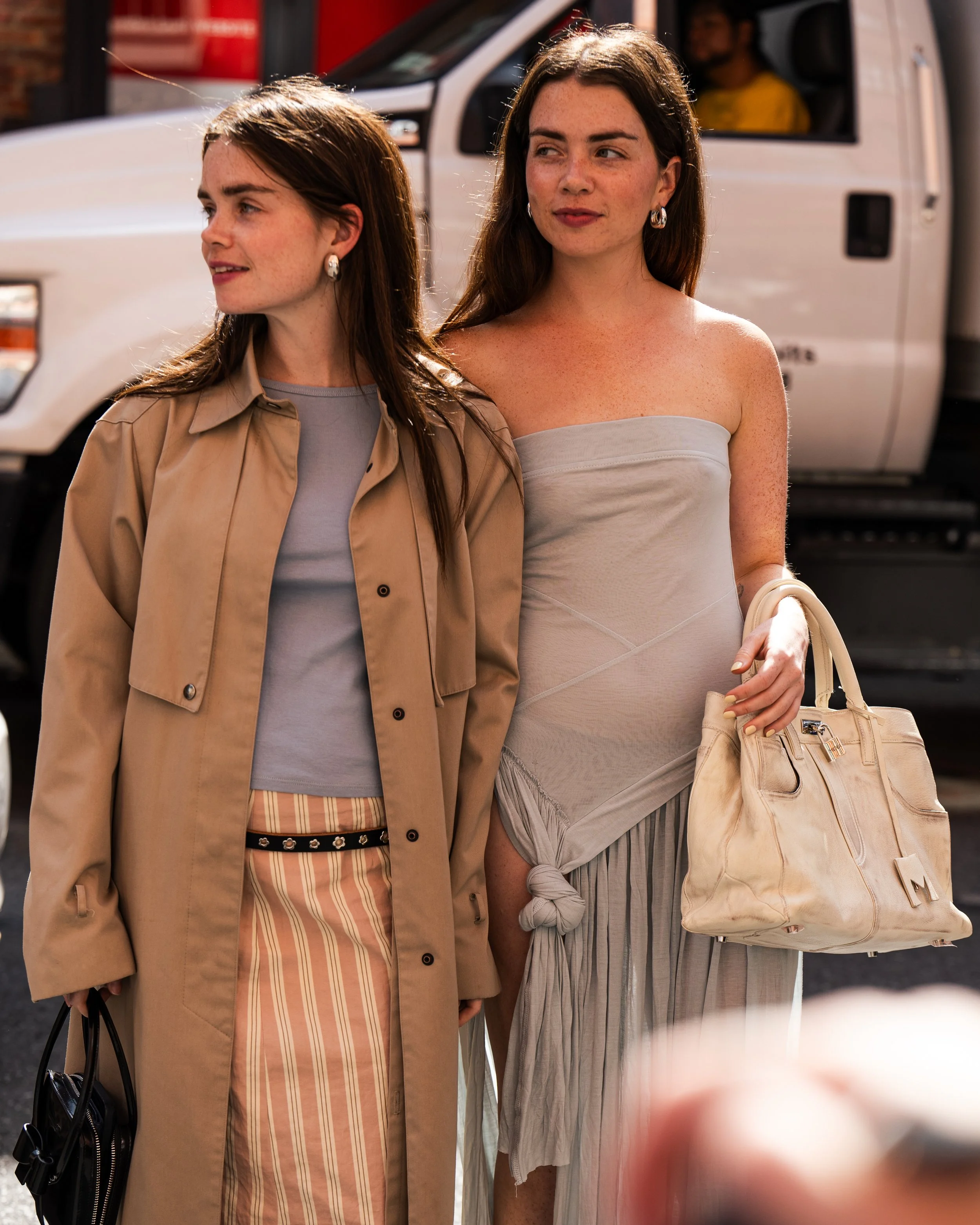 Two women standing on the street with a white vehicle in the background. One woman is wearing a tan trench coat over a gray top and striped pants, holding a black handbag. The other woman is wearing a strapless beige dress, holding a beige handbag, and has a confident expression.