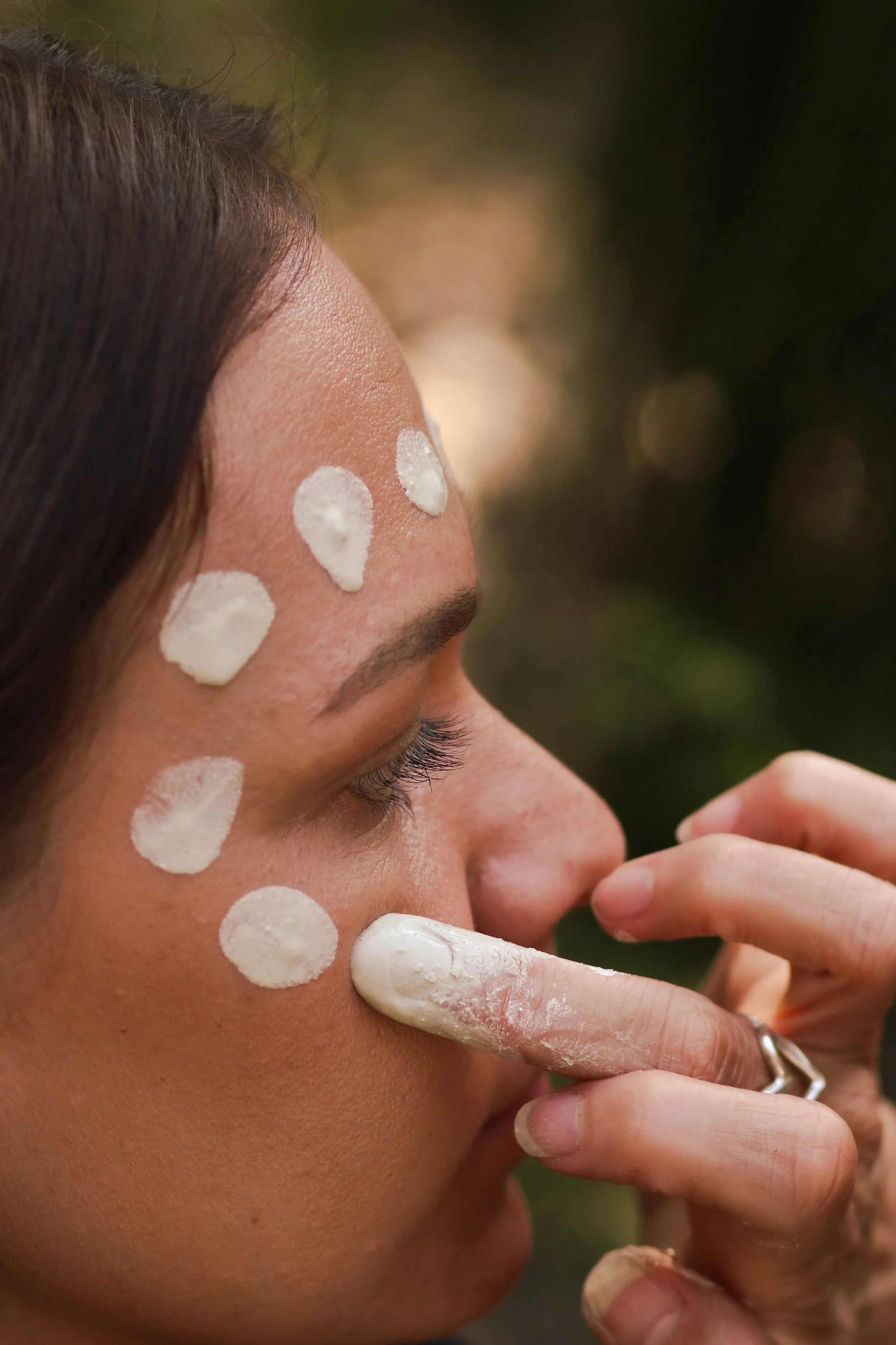 A woman applies white cream or lotion to her face, with several spots of the substance visible on her forehead, cheek, and near her nose, against a blurred outdoor background.