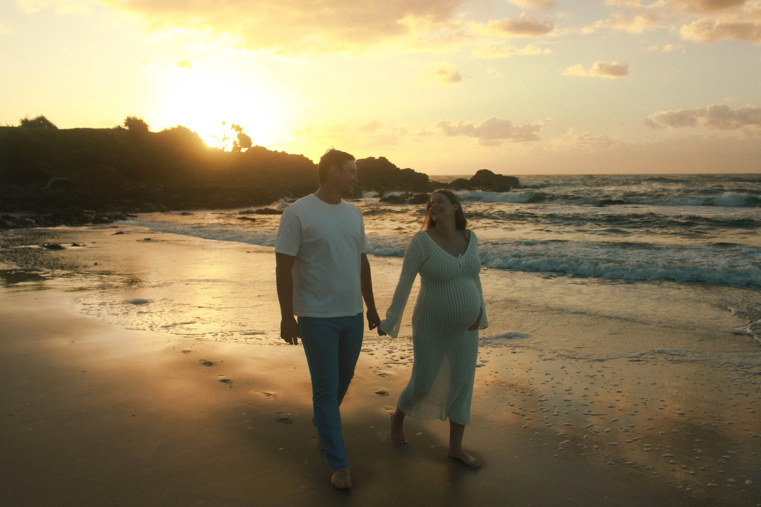 A pregnant woman and a man walking hand in hand on the beach at sunset, smiling and looking at each other.