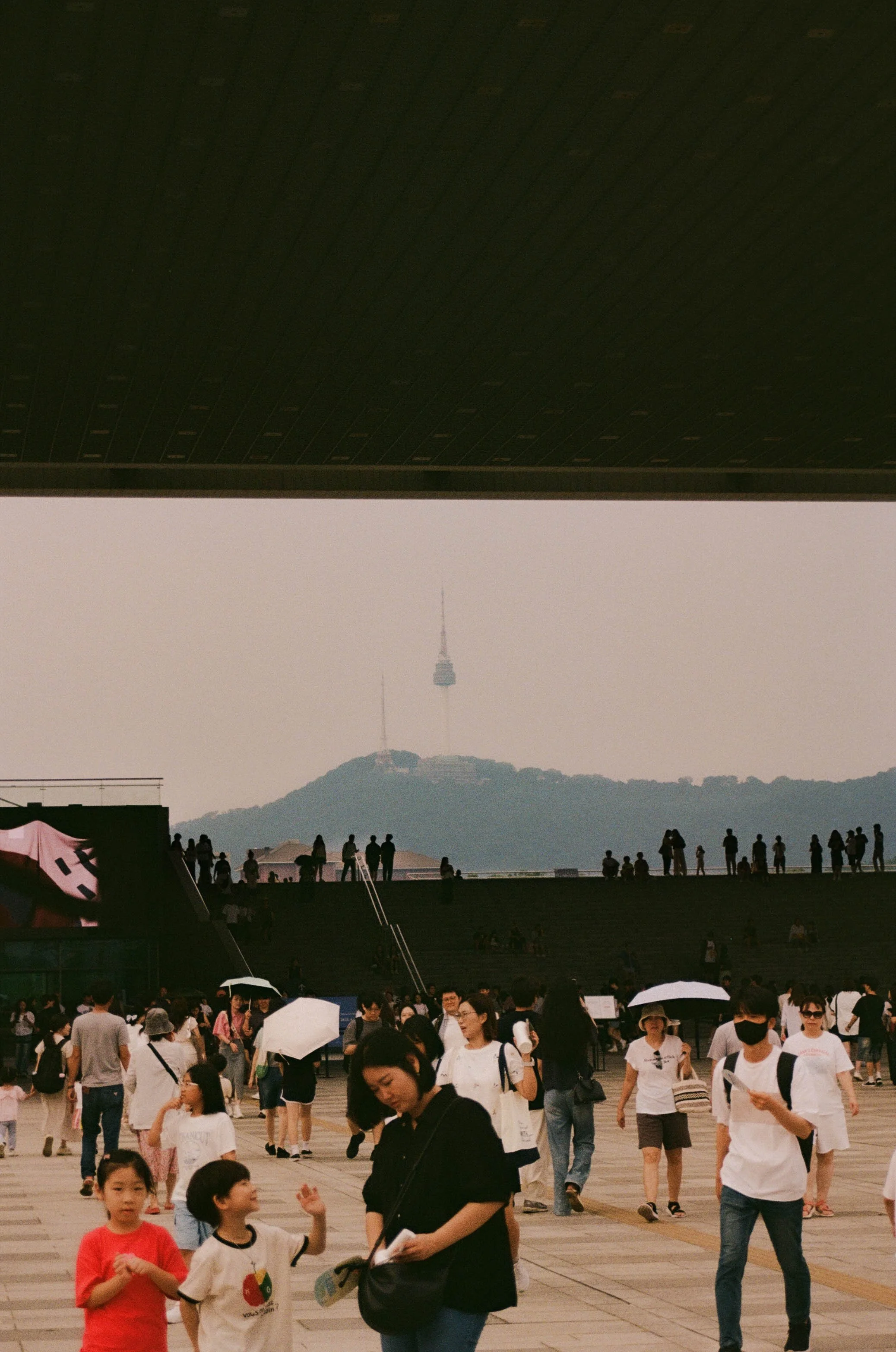 People gathering at an outdoor venue in Sout Korea with a hill and communication tower in the background, some holding umbrellas.