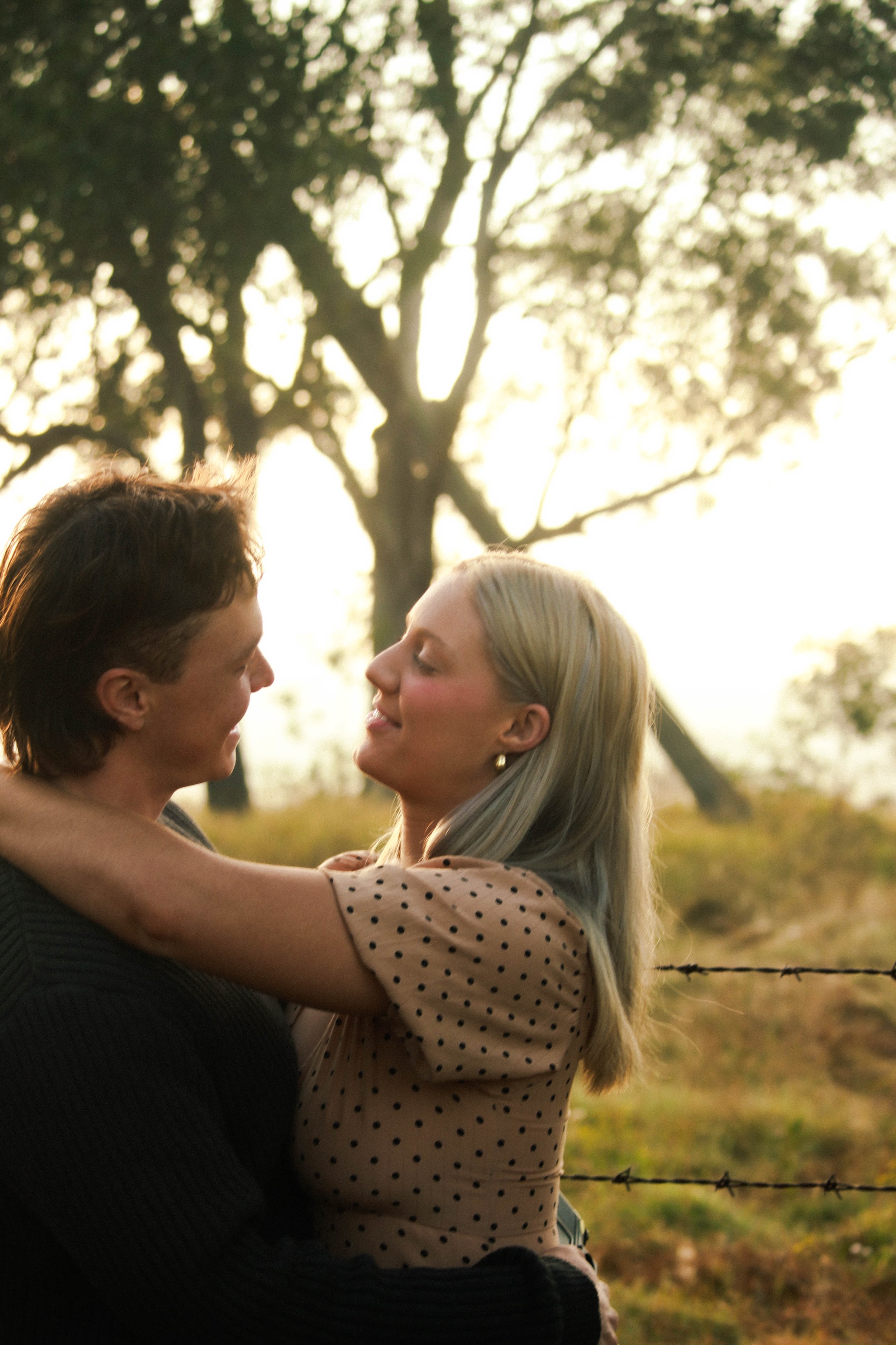 A couple embracing outdoors at sunset, with trees and a barbed wire fence in the background.