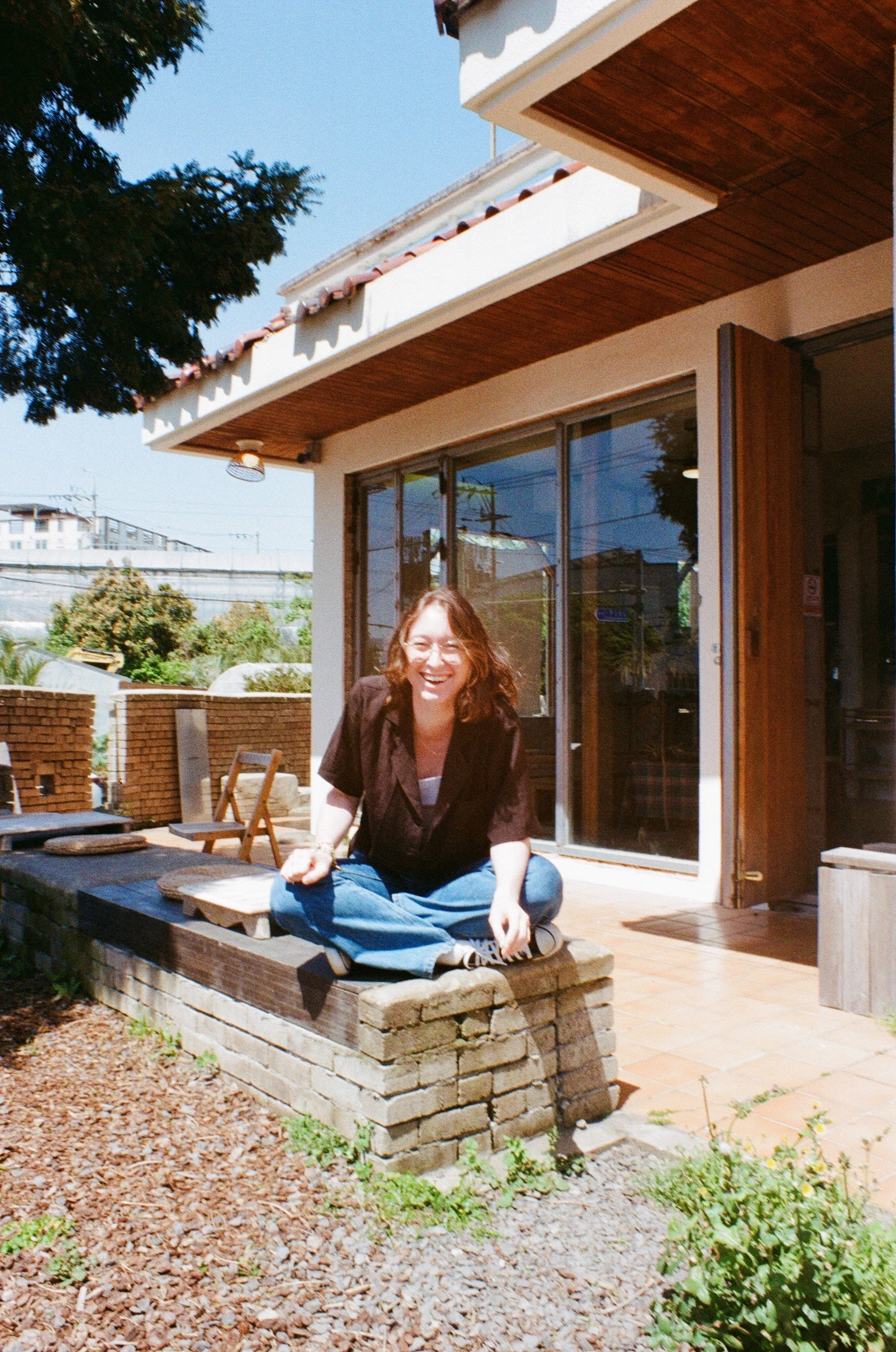 A woman sitting cross-legged on a brick and wood ledge outside a building, smiling, with a patio and chairs in the background.
