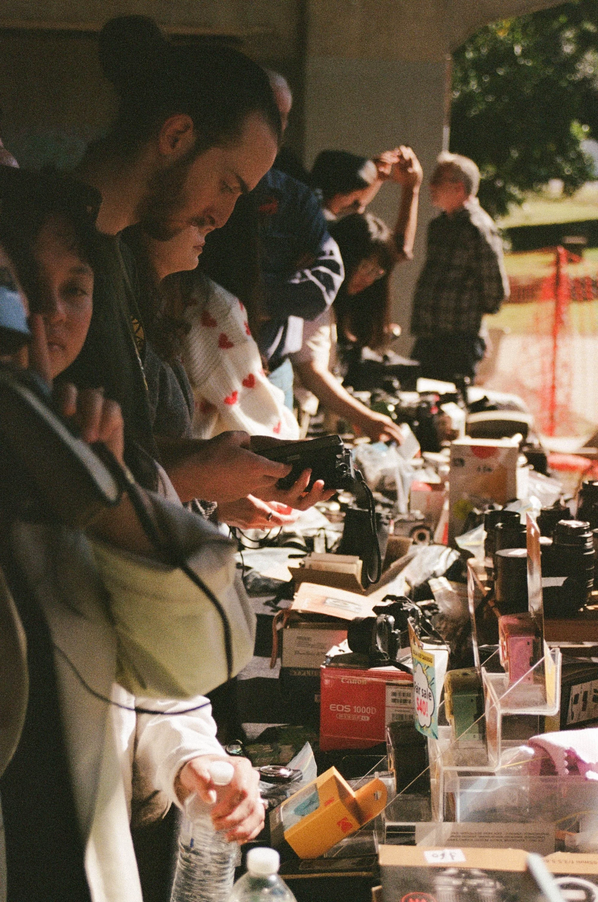 People browsing cameras and electronics at outdoor market table, some examining items and others looking at display