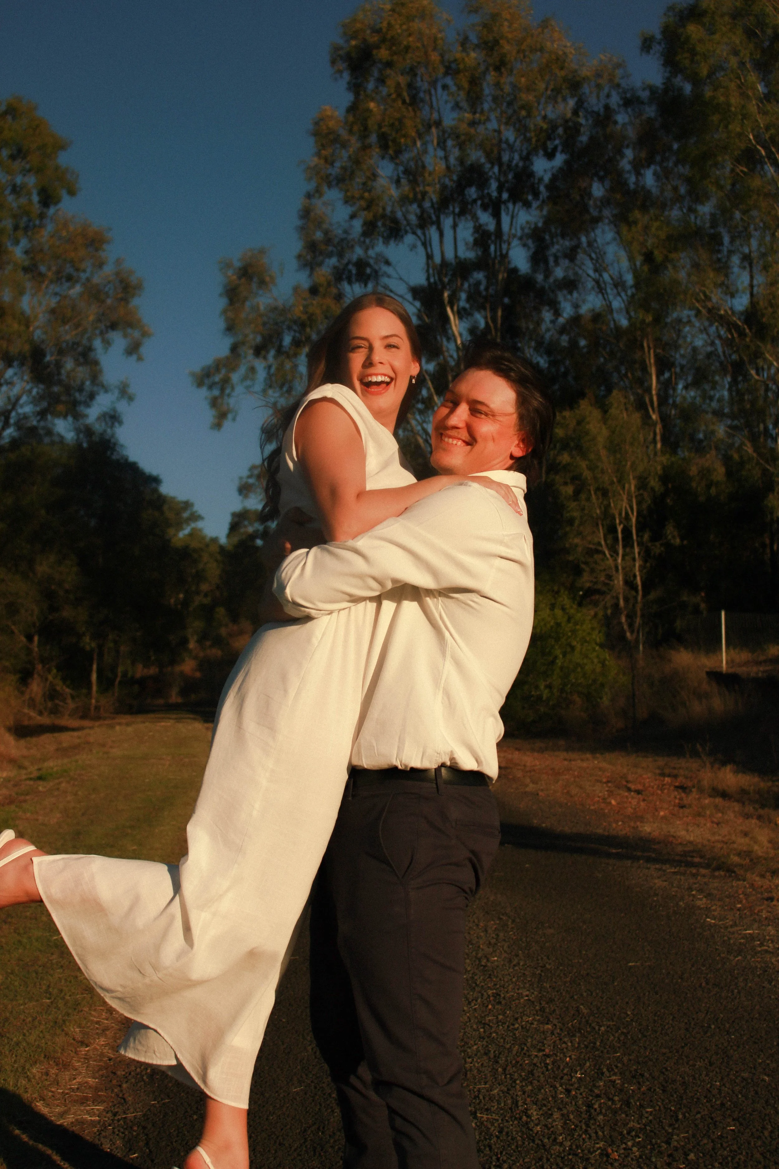 A man lifting a woman in a happy, outdoor setting during sunset, with trees and a clear sky in the background.