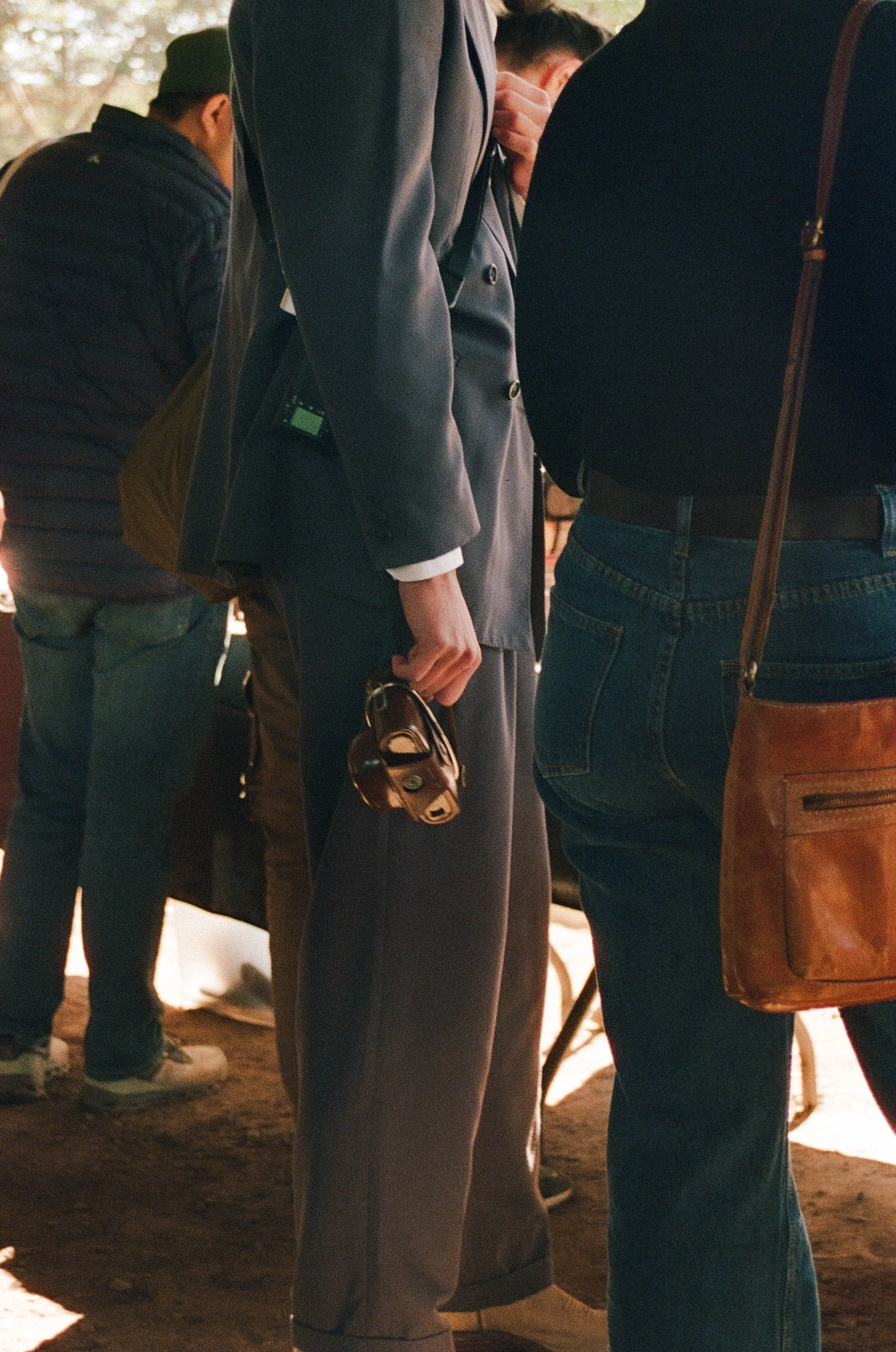 Several people standing close together, seen from the waist down. One person is holding a small, brown handbag. Others have backpacks or bags, and they are dressed in casual and business attire.