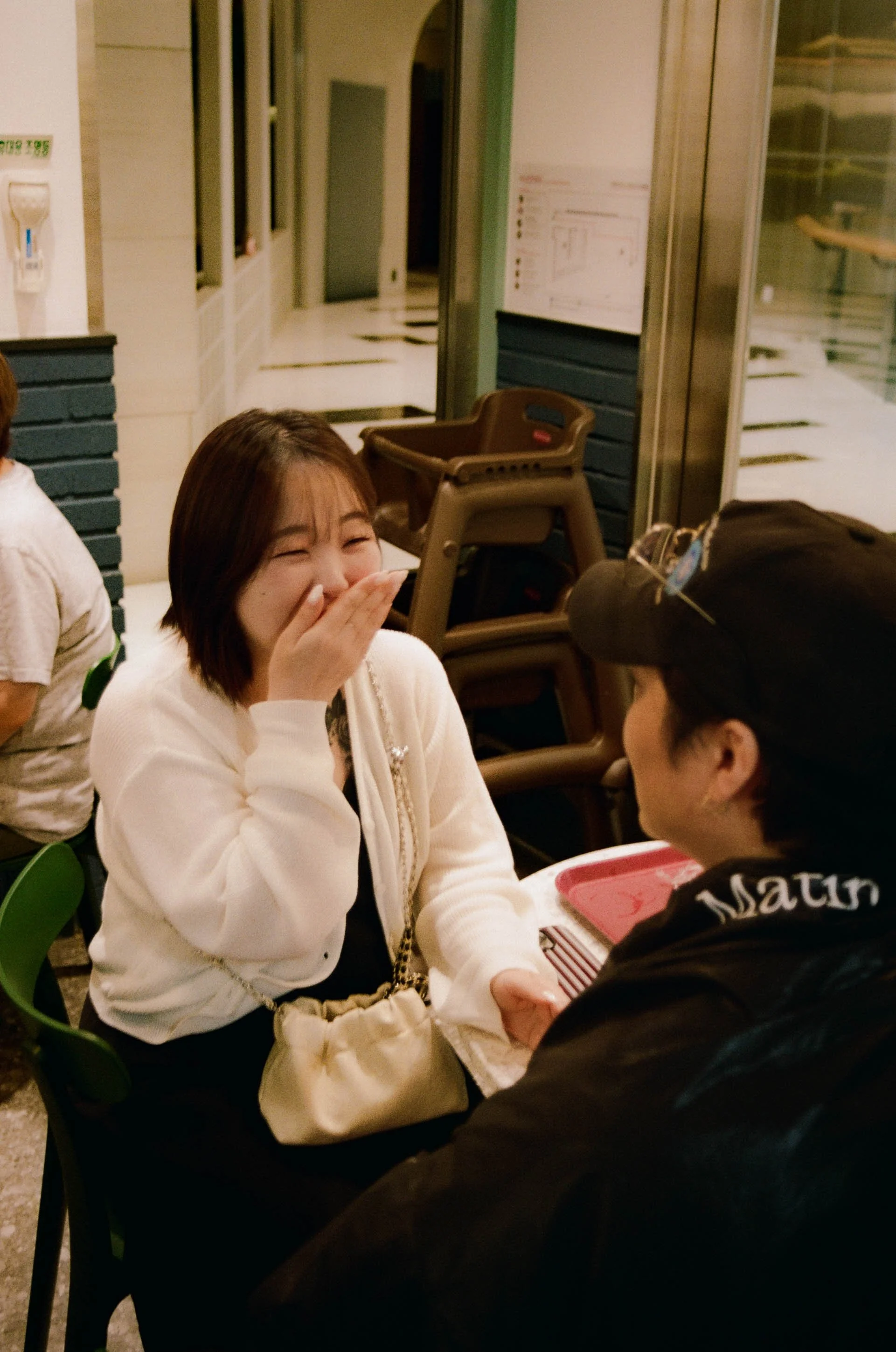 Two women sitting across from each other, smiling and laughing, inside a restaurant. One woman with short dark hair is covering her mouth in amusement, wearing a white cardigan, while the other woman with glasses and a black cap is facing her, holding a pink tray.