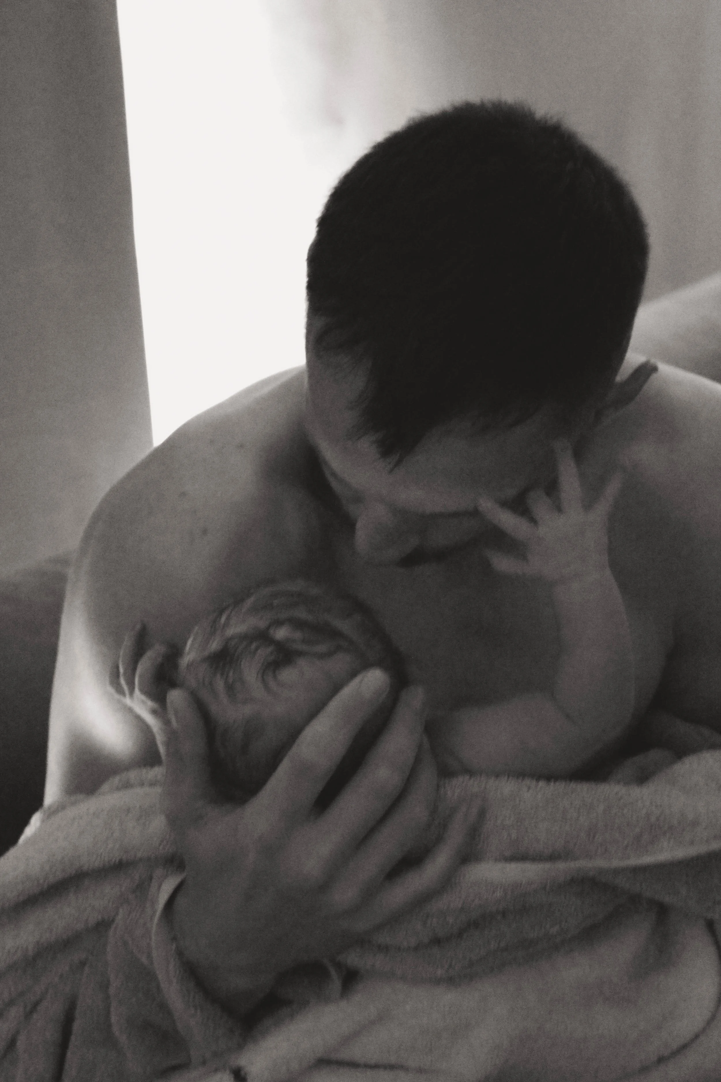 A man holding a newborn baby against his chest, covering part of his face with his hand, in a black and white photo.