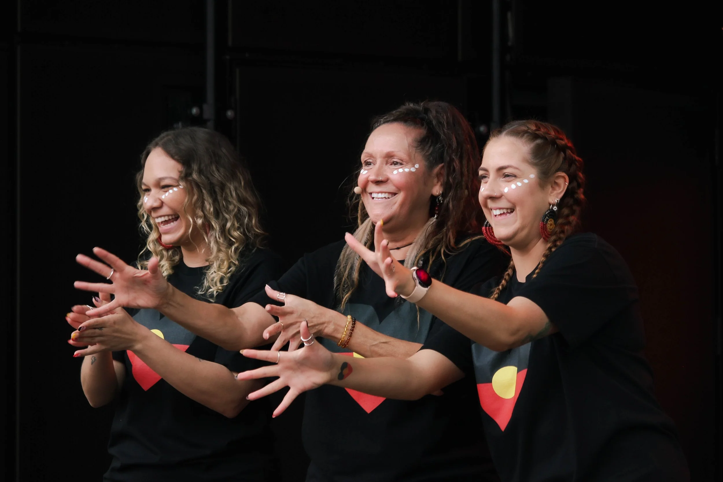 Three women with face paint and coordinated black T-shirts, smiling and gesturing with their hands during a group activity indoors.