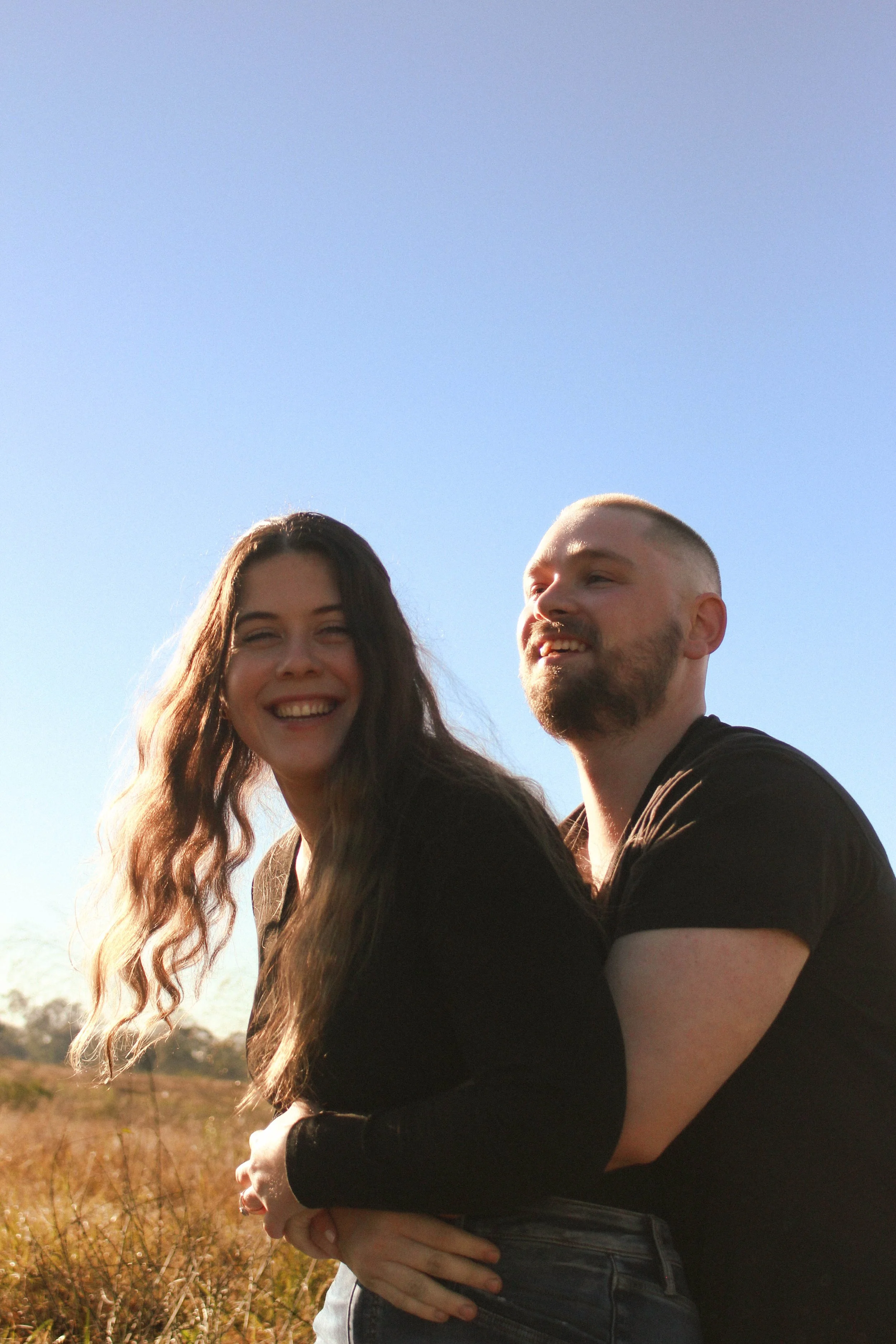 A young couple outdoors in a field, smiling and enjoying a sunny day with a clear blue sky.