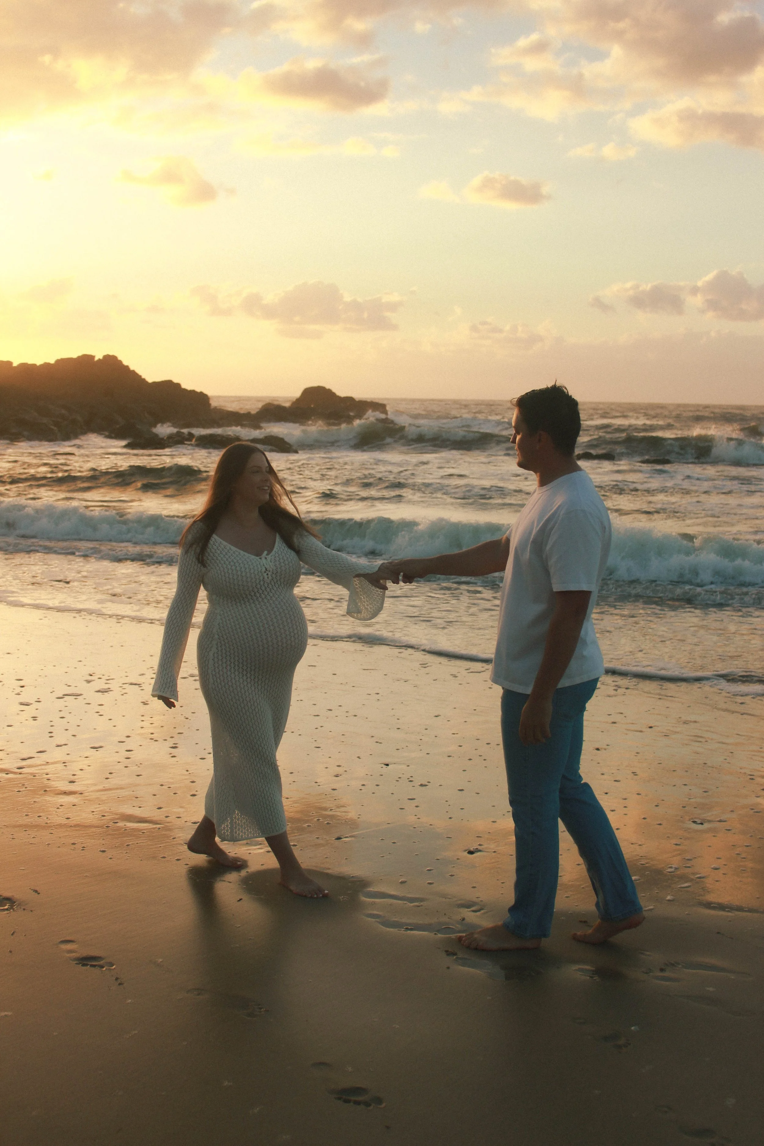 A couple holding hands on the beach at sunset with the ocean waves and rocky shoreline in the background.