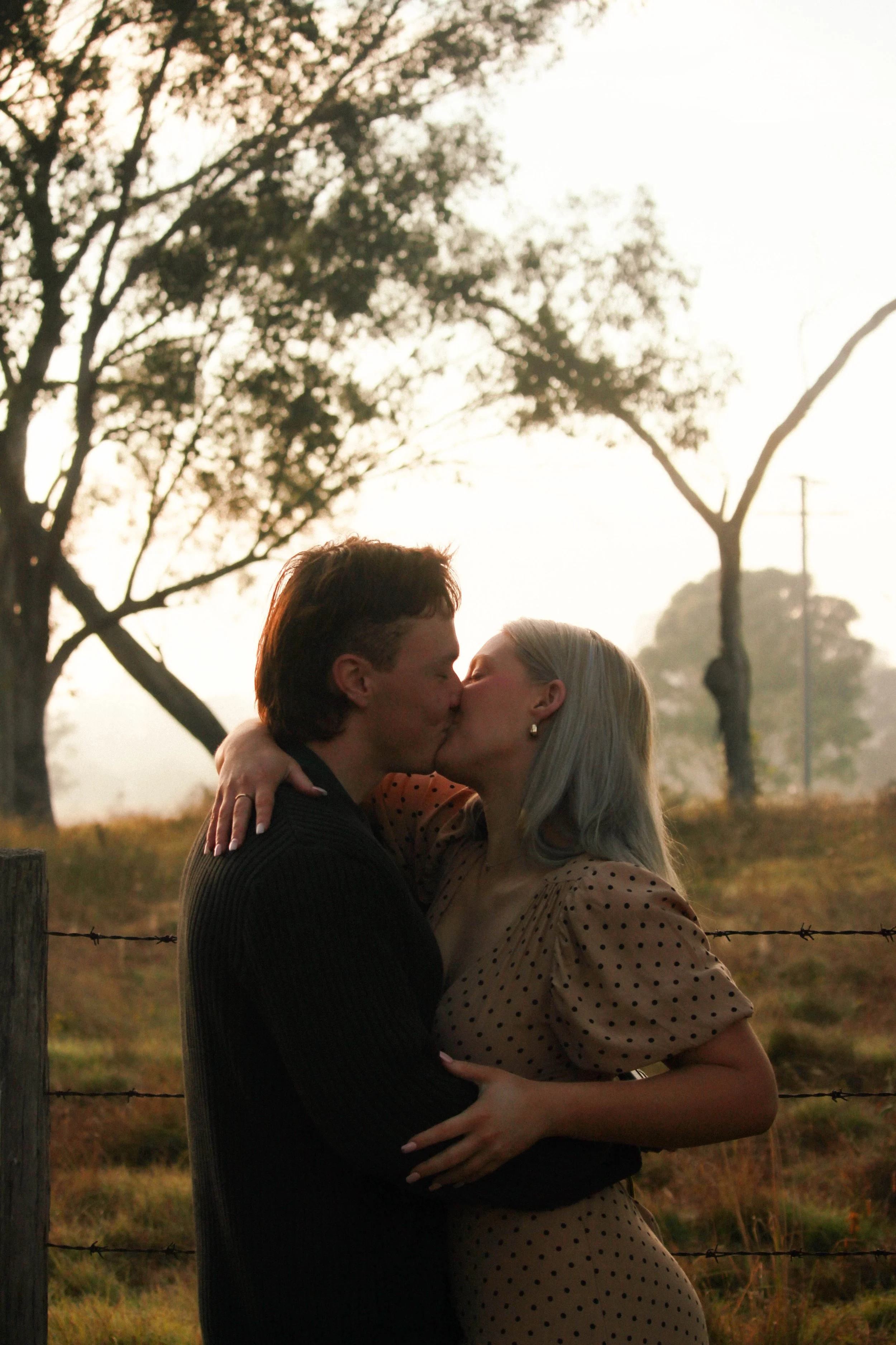 A couple sharing a kiss outdoors near a barbed wire fence with trees and a sunset in the background.