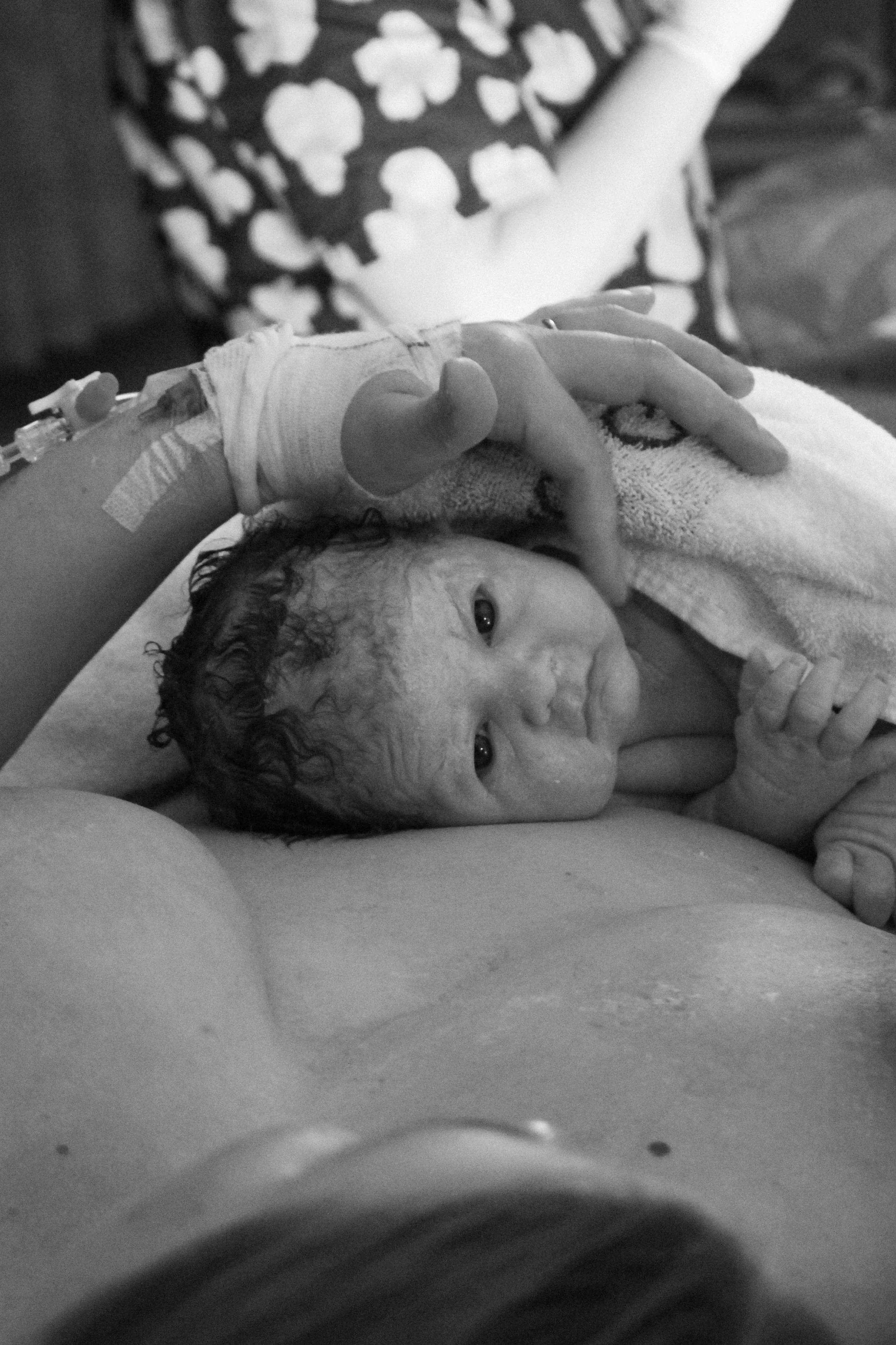 Newborn baby with wet hair lying on an adult's chest, with an IV in the baby's hand, in a black and white photograph.