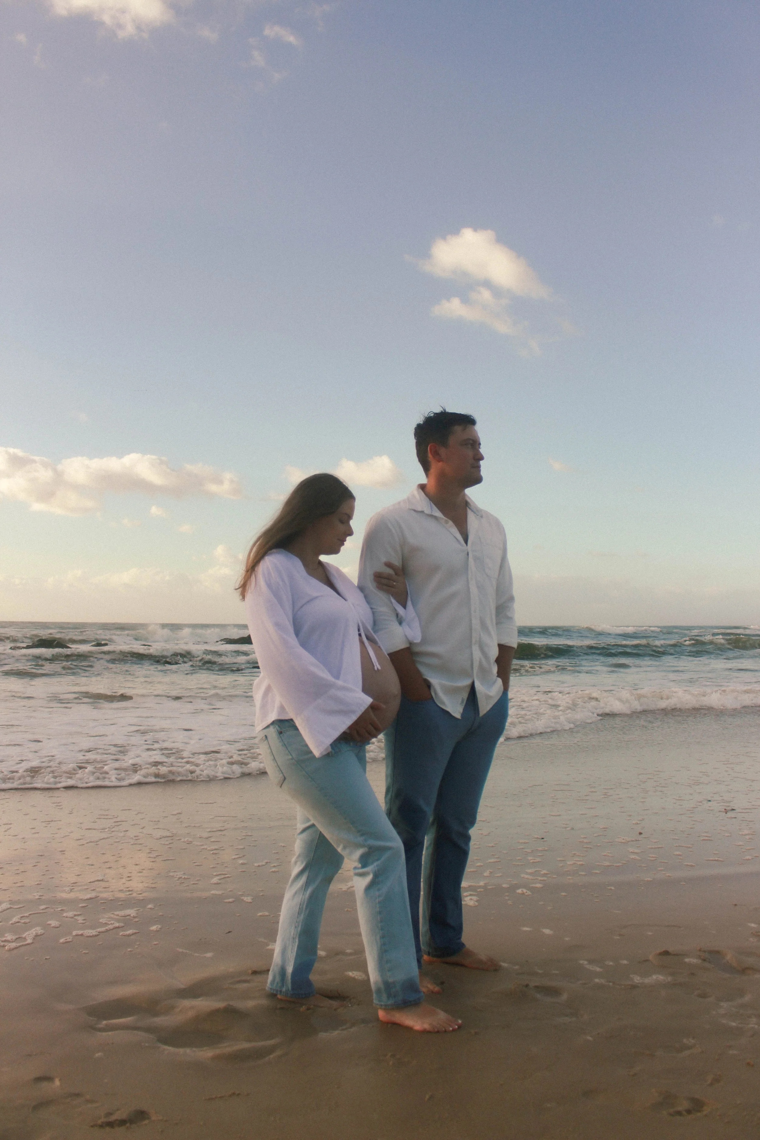 A pregnant woman and a man standing on the beach near the ocean, with the woman placing her hand on her baby bump. The woman has long hair and is wearing a white blouse and light blue jeans. The man has short hair and is wearing a white shirt and blue jeans, both barefoot. The sky is partly cloudy.