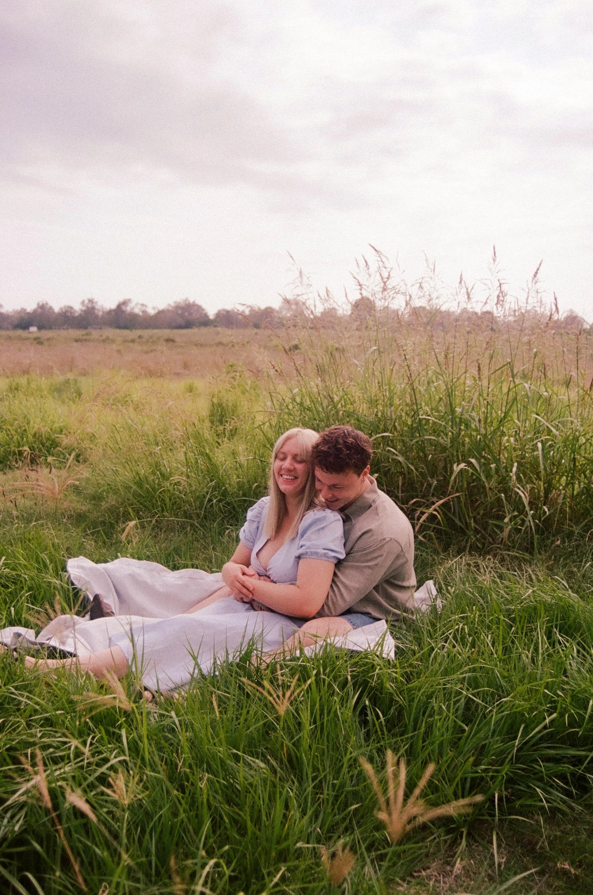 A couple sitting on a blanket in a grassy field, embracing and smiling happily.