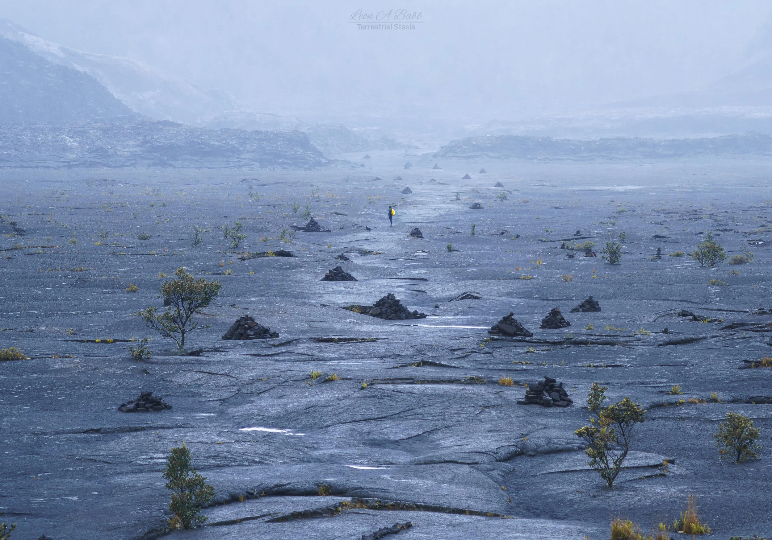A person wearing a yellow jacket walking through a vast, foggy volcanic landscape with small rocks and sparse, low vegetation.