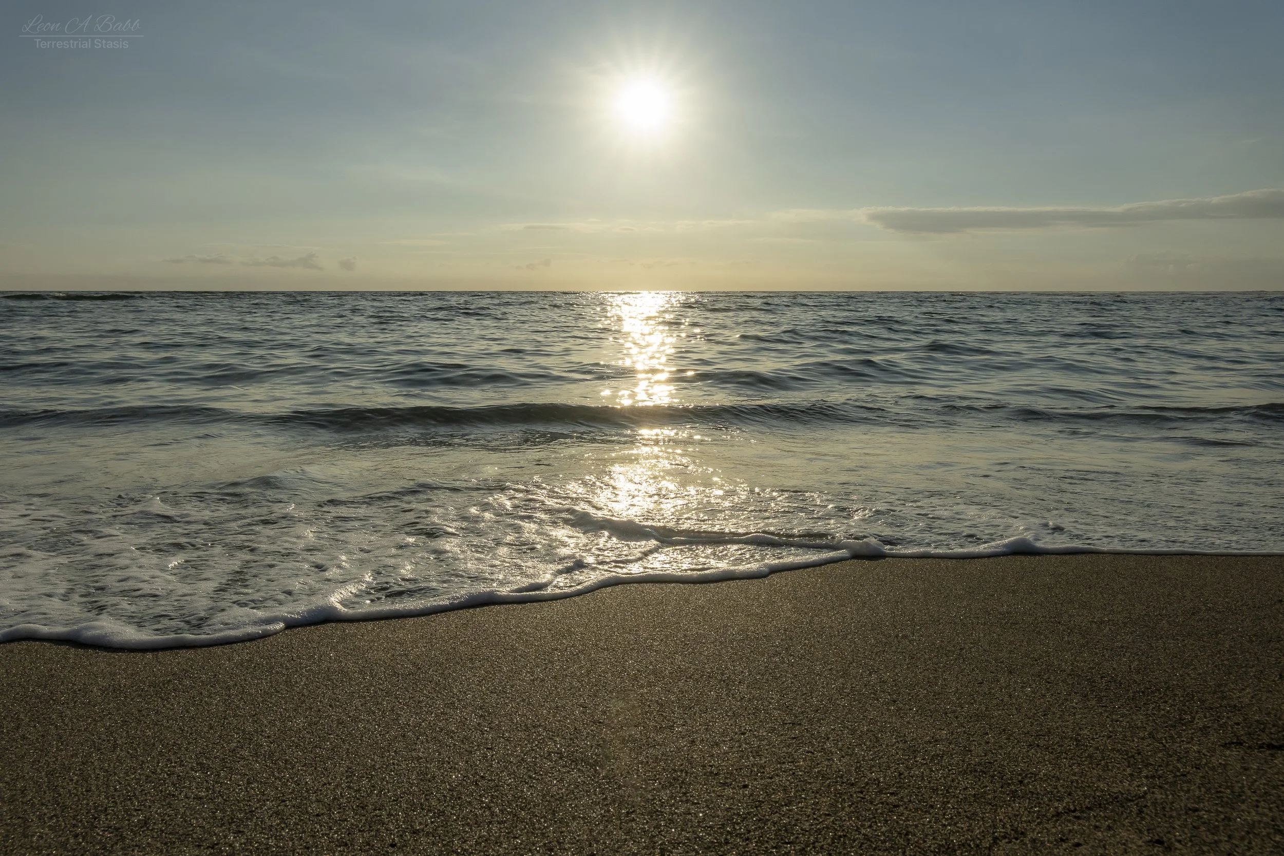 Photo of a beach during sunset with the sun low in the sky, gentle waves rolling onto the sandy shore, and a partly cloudy sky.