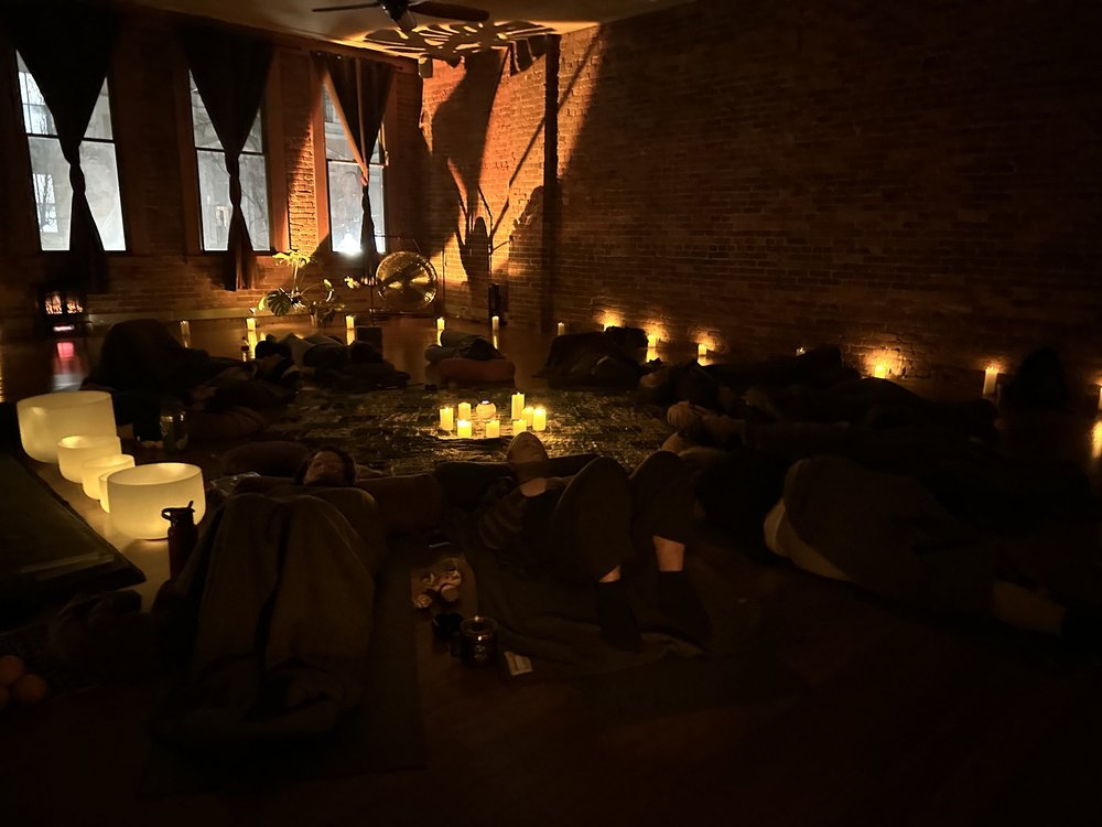 A candlelit Aloka sound bath at Bodha Yoga: brick walls, tall windows, crystal bowls glowing on the floor, people resting under blankets