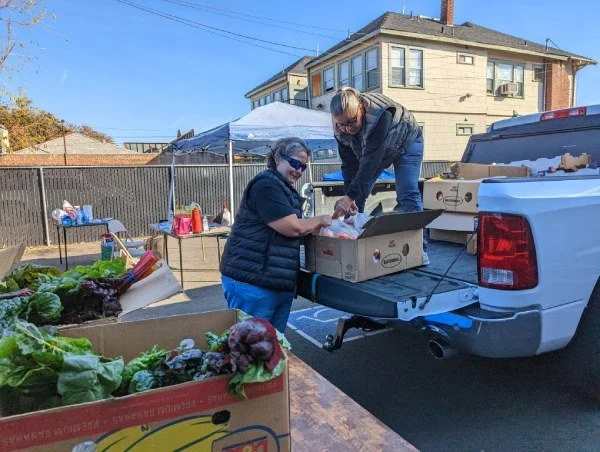 Two WFWA volunteers work together to unload boxes of fruits and vegetables from the bed of a pickup truck. Boxes filled with fresh green produce line a table in the foreground