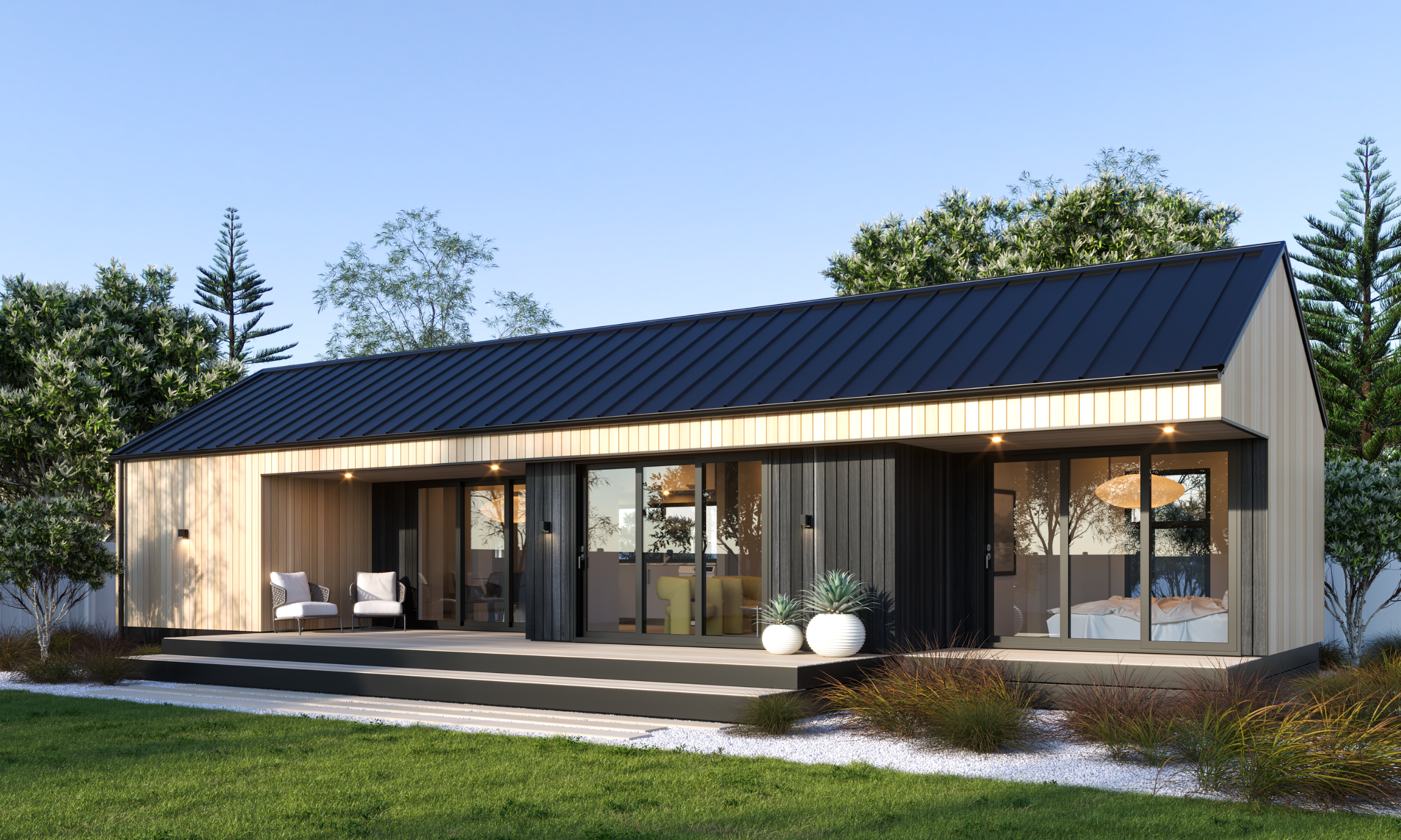 Modern house with black metal roof, large glass sliding doors, outdoor seating, surrounded by greenery and trees at dusk.