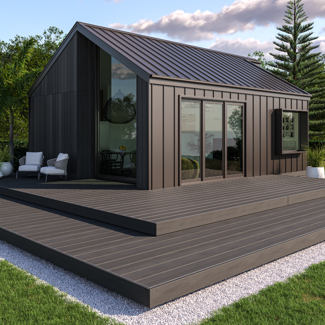 Modern black house with large glass sliding doors on a wooden deck, outdoor white seating, surrounded by greenery and a blue sky with clouds.