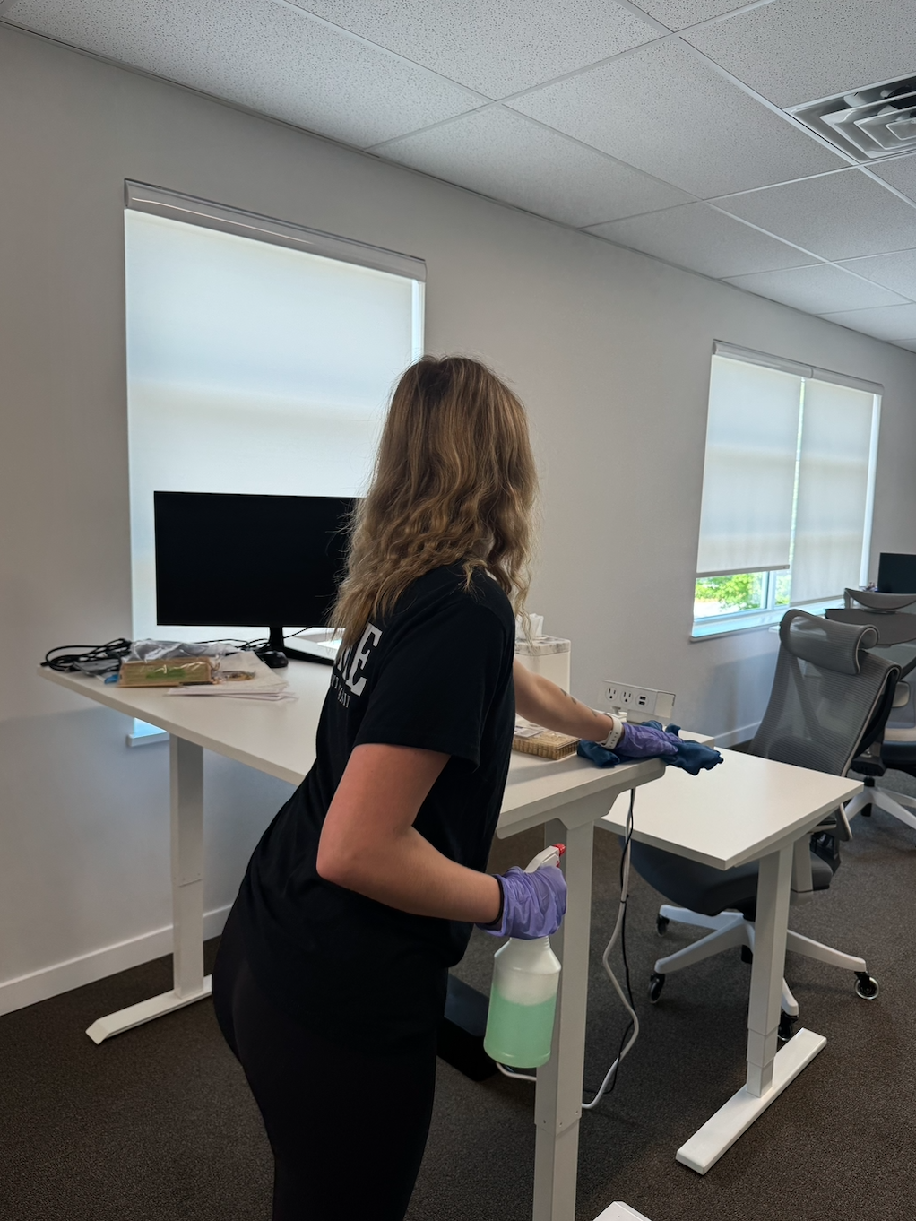 A woman cleaning a white office desk with a spray bottle, wearing purple gloves in a modern office with white walls and shades partially drawn over windows.