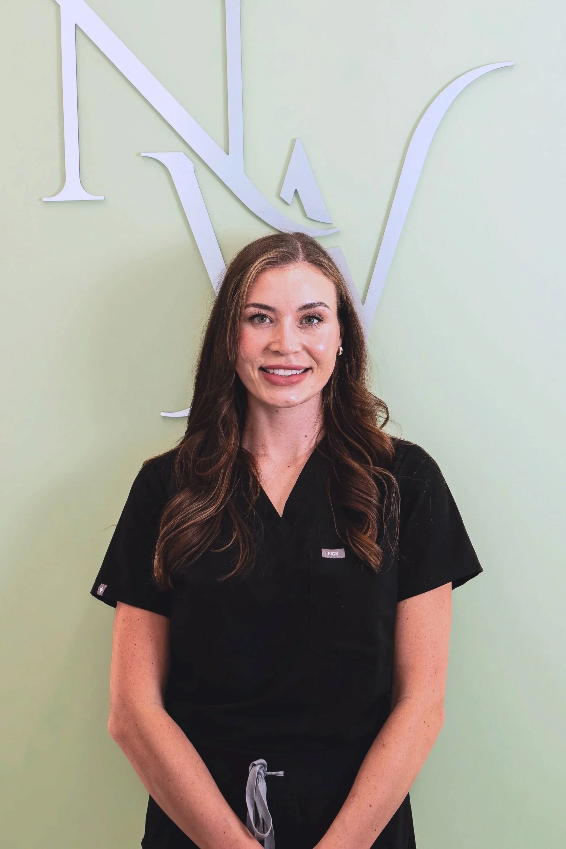 A woman with long wavy brown hair, wearing black scrubs, standing in front of a light green wall with a large white logo, for Nova Wellness, behind her.