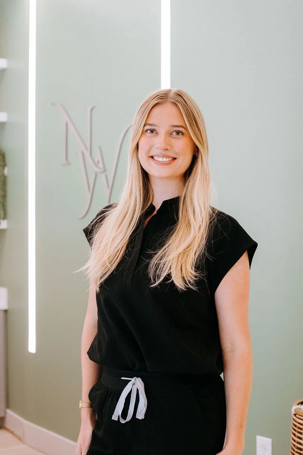 A young woman with long blonde hair smiling, wearing a black short-sleeve top and black pants with a gray drawstring, standing in front of a light green wall with a decorative logo for Nova Wellness.