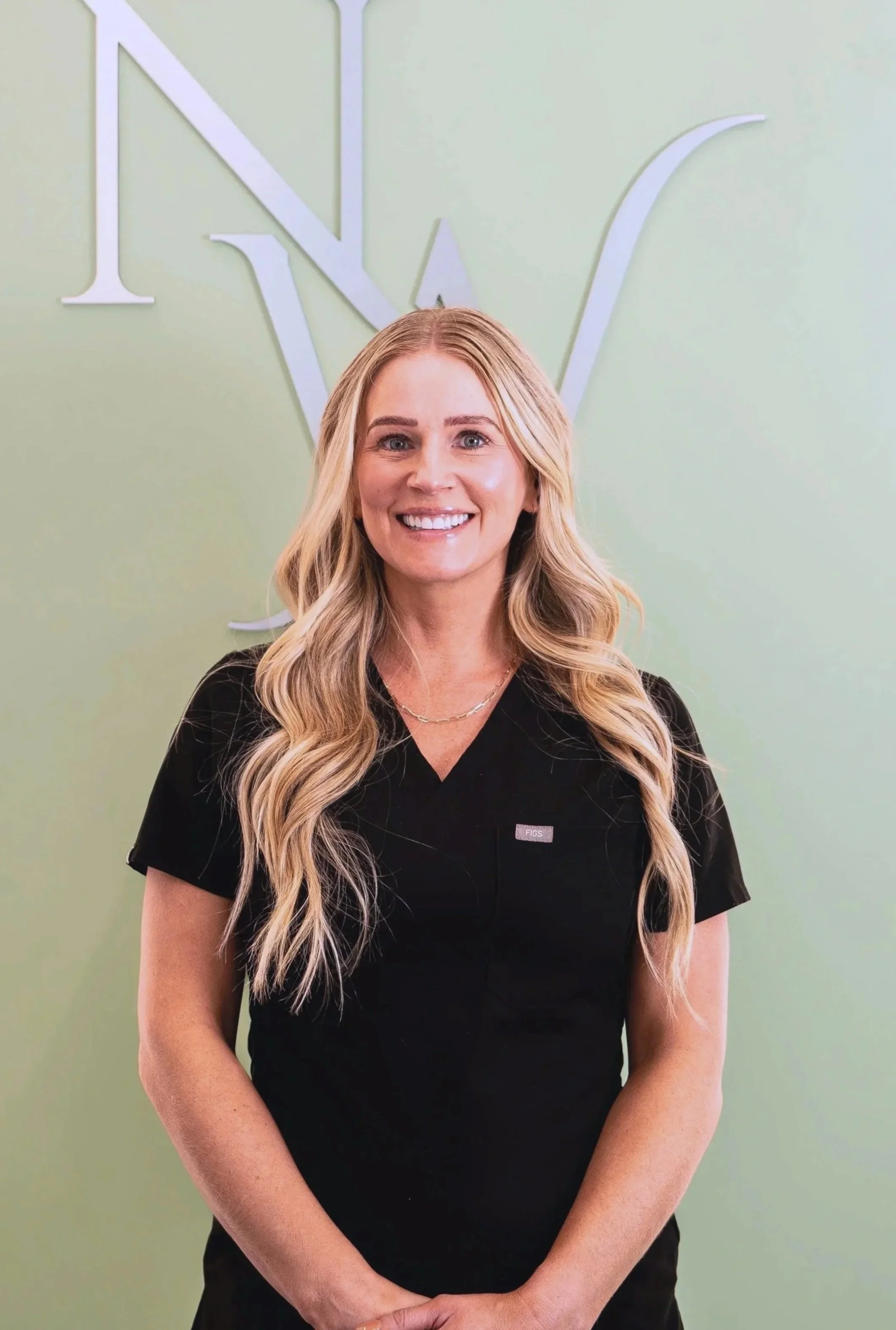 A blonde woman smiling in a black medical uniform standing in front of a pale green wall with a large Nova Wellness logo.