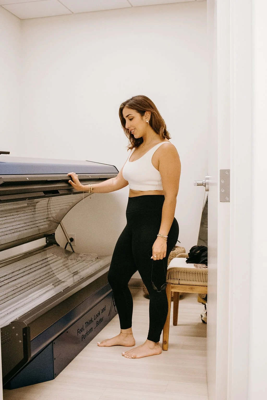A woman standing next to a light therapy bed at Nova Wellness, barefoot, in a room with beige walls and a wooden floor, smiling and looking down.