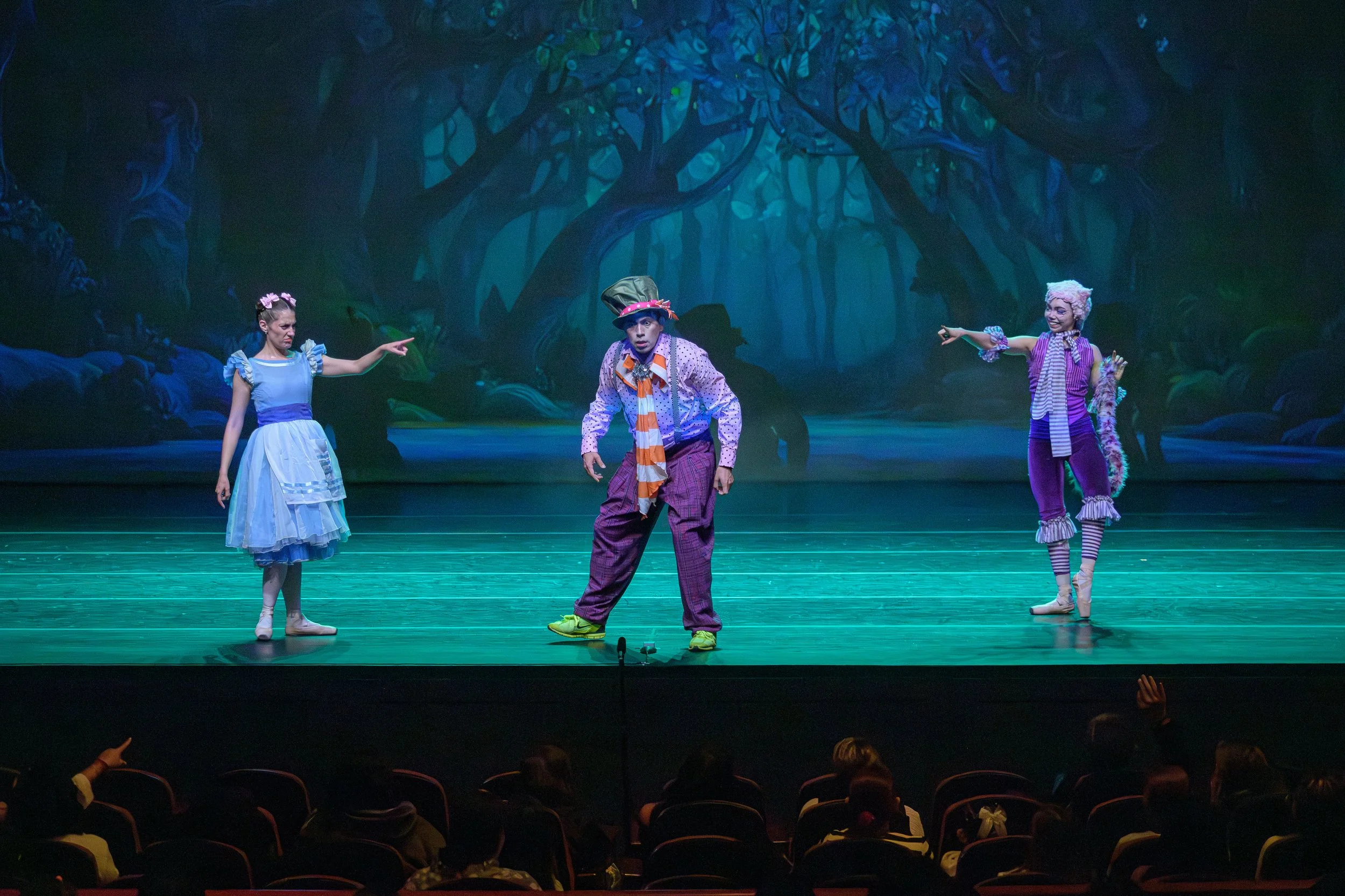Stage scene from a theatrical performance with three actors in colorful costumes, set against a dark, fantasy forest backdrop with trees and foliage.