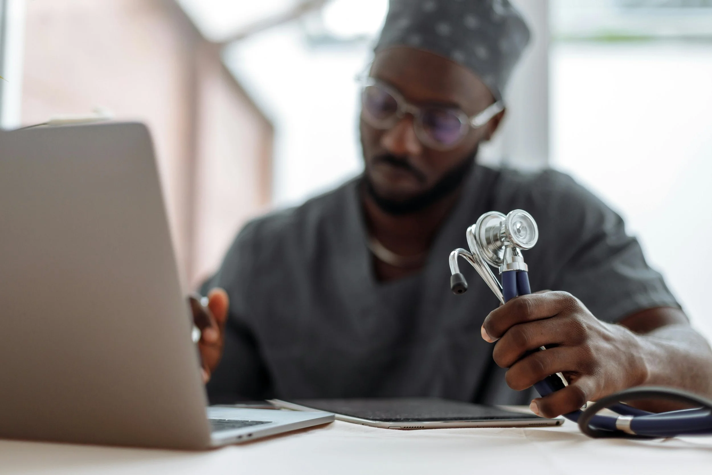 Man sits at laptop. He is wearing scrubs, and holds a stethoscope in one hand.