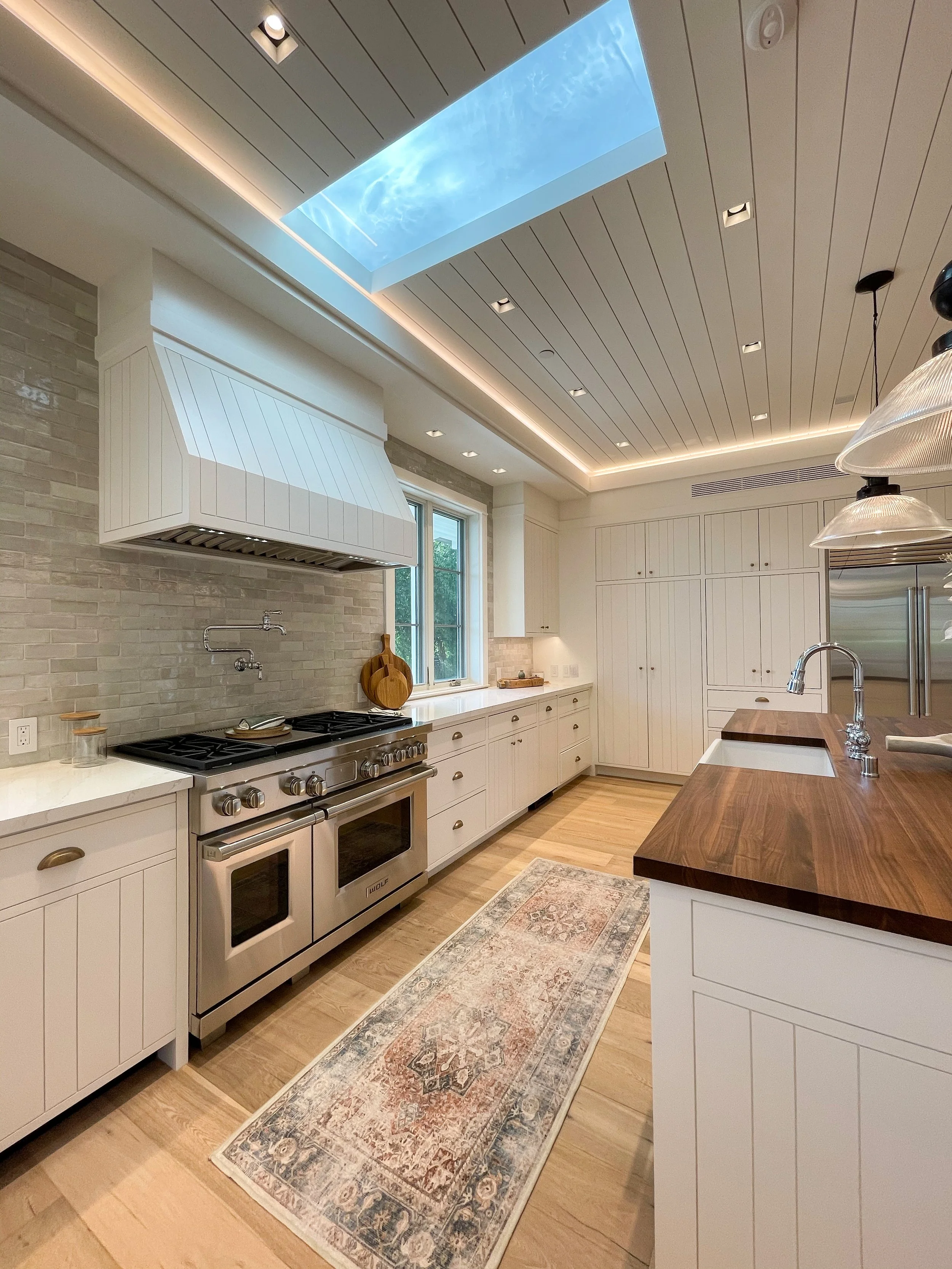 Modern kitchen with white cabinetry, a stainless steel stove, and a wooden kitchen island. Ceiling has a skylight showing a blue sky with clouds.