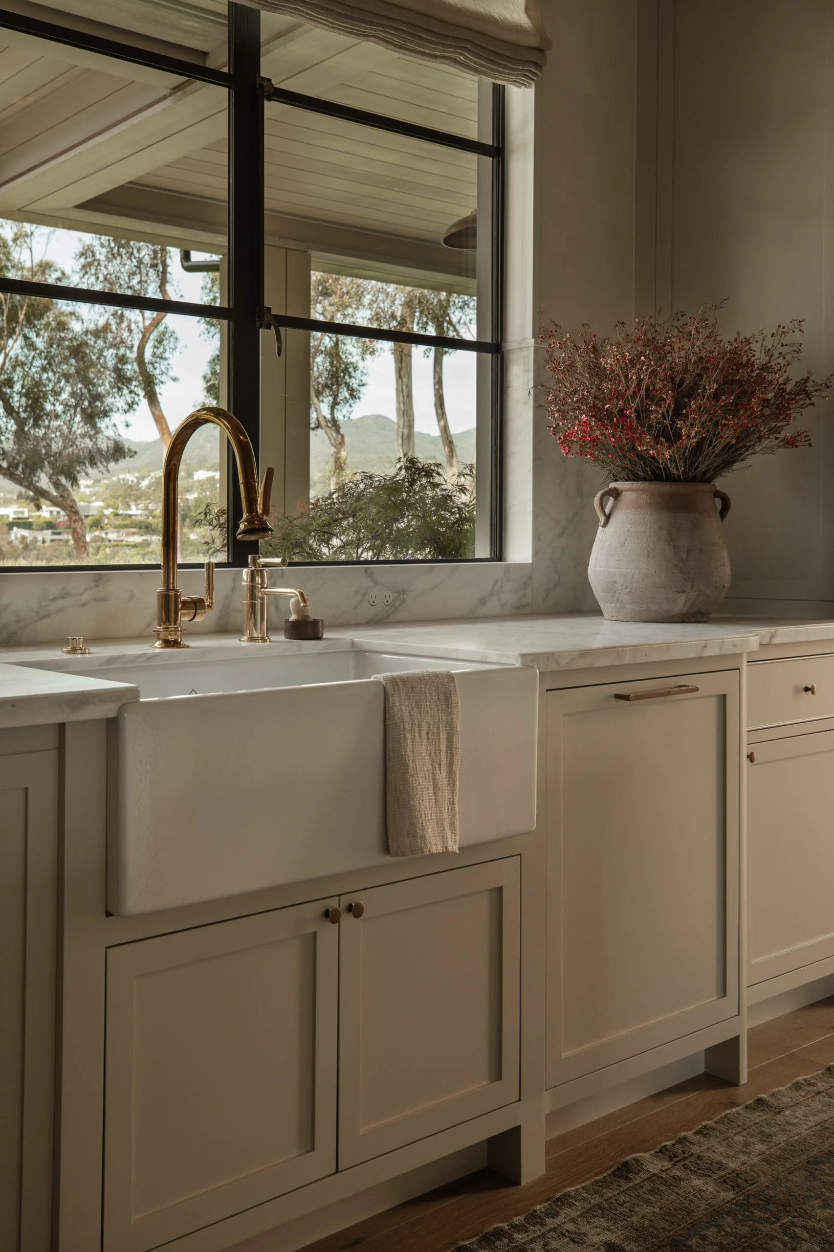 Kitchen with white cabinetry, marble countertops, a farmhouse sink, a brass faucet, a large window with a view of trees and mountains, and a beige vase with pink and red dried flowers.