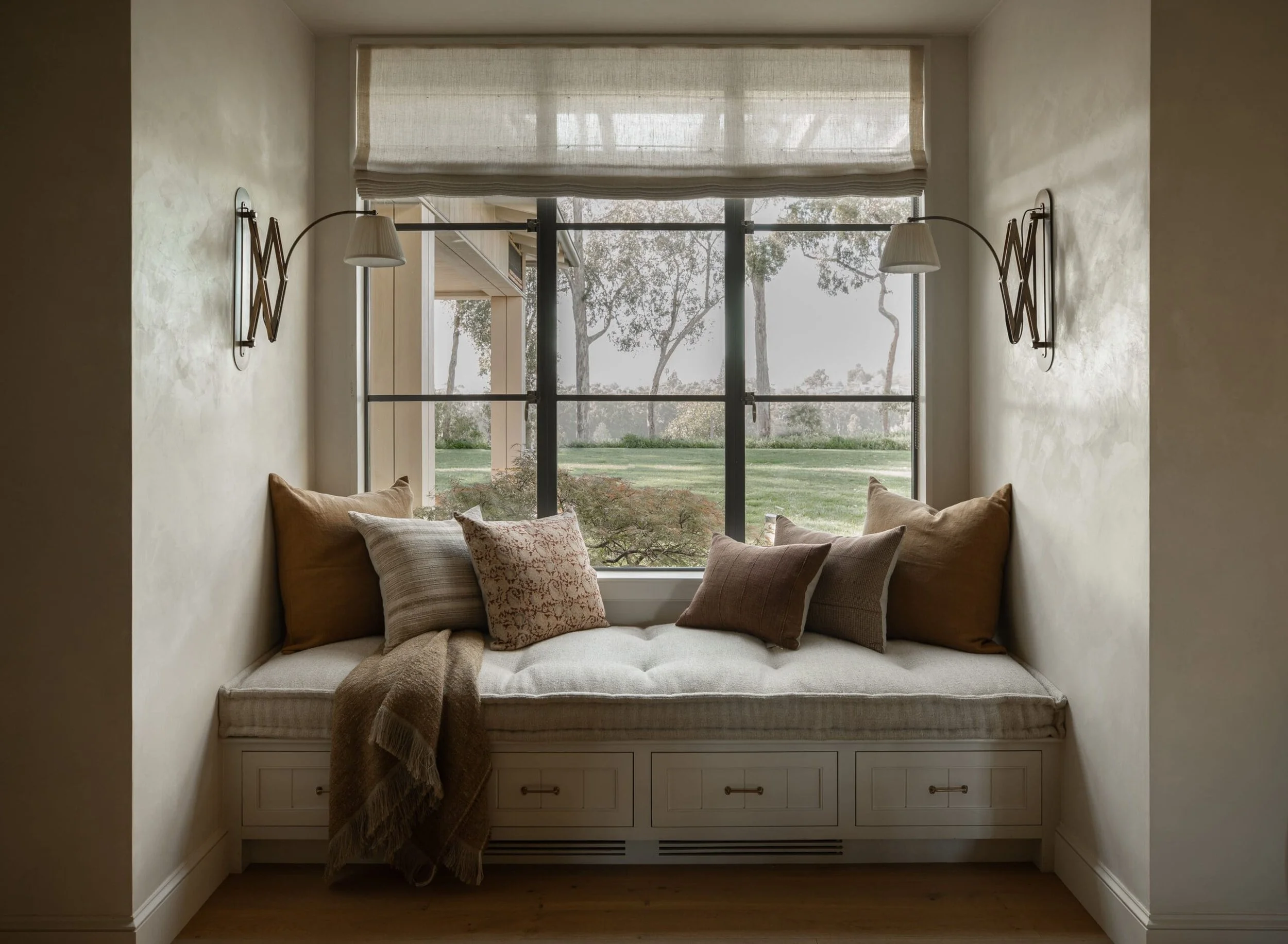 A cozy window nook with a cushioned bench and multiple throw pillows in neutral tones, flanked by wall-mounted reading lamps, overlooking a lush outdoor landscape through large glass windows.