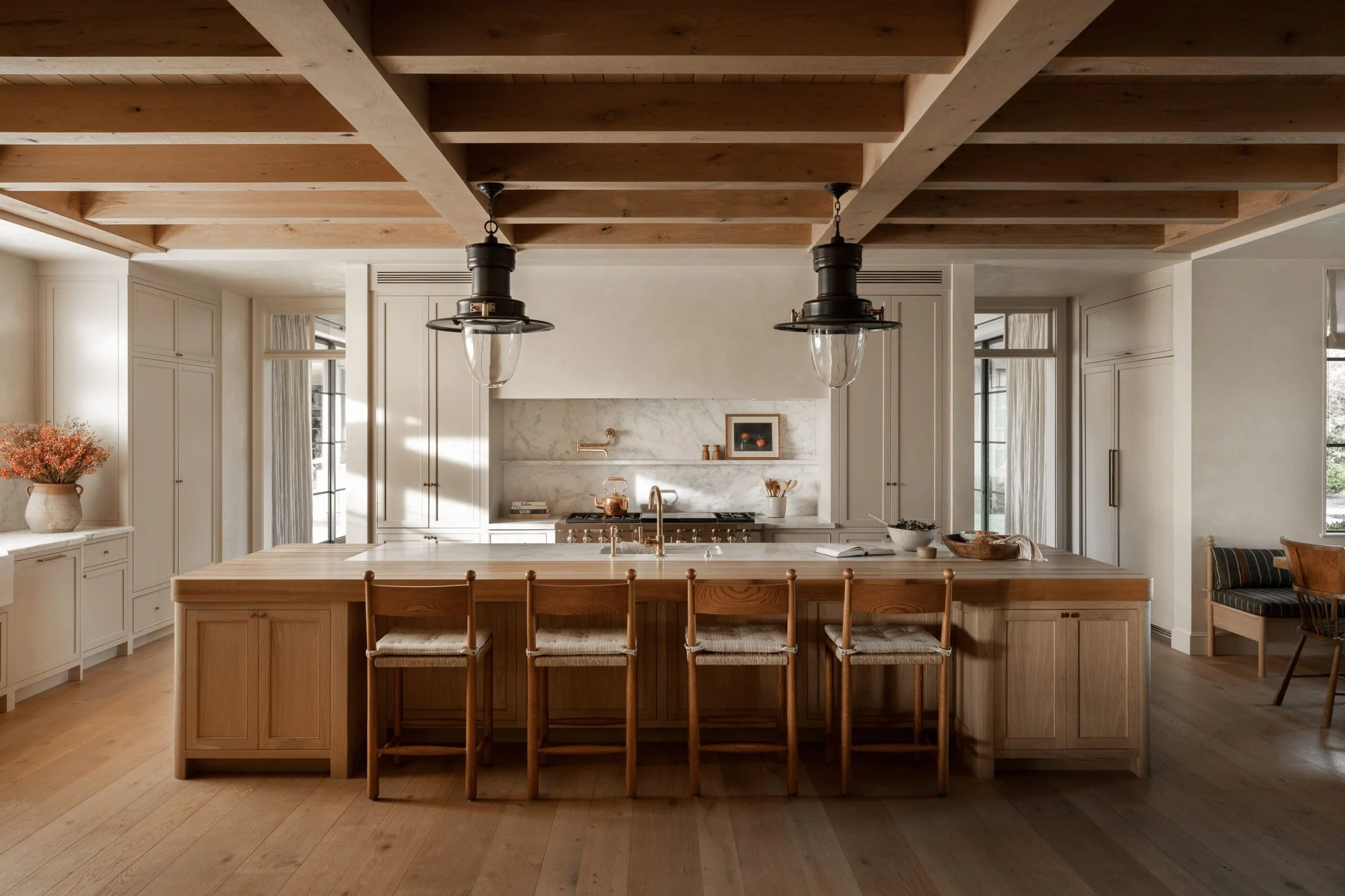A spacious modern kitchen with a large wooden island counter, four wooden chairs with woven seats, white cabinetry, marble backsplash, and two black pendant lights hanging from a wooden ceiling. Light streams in through windows.