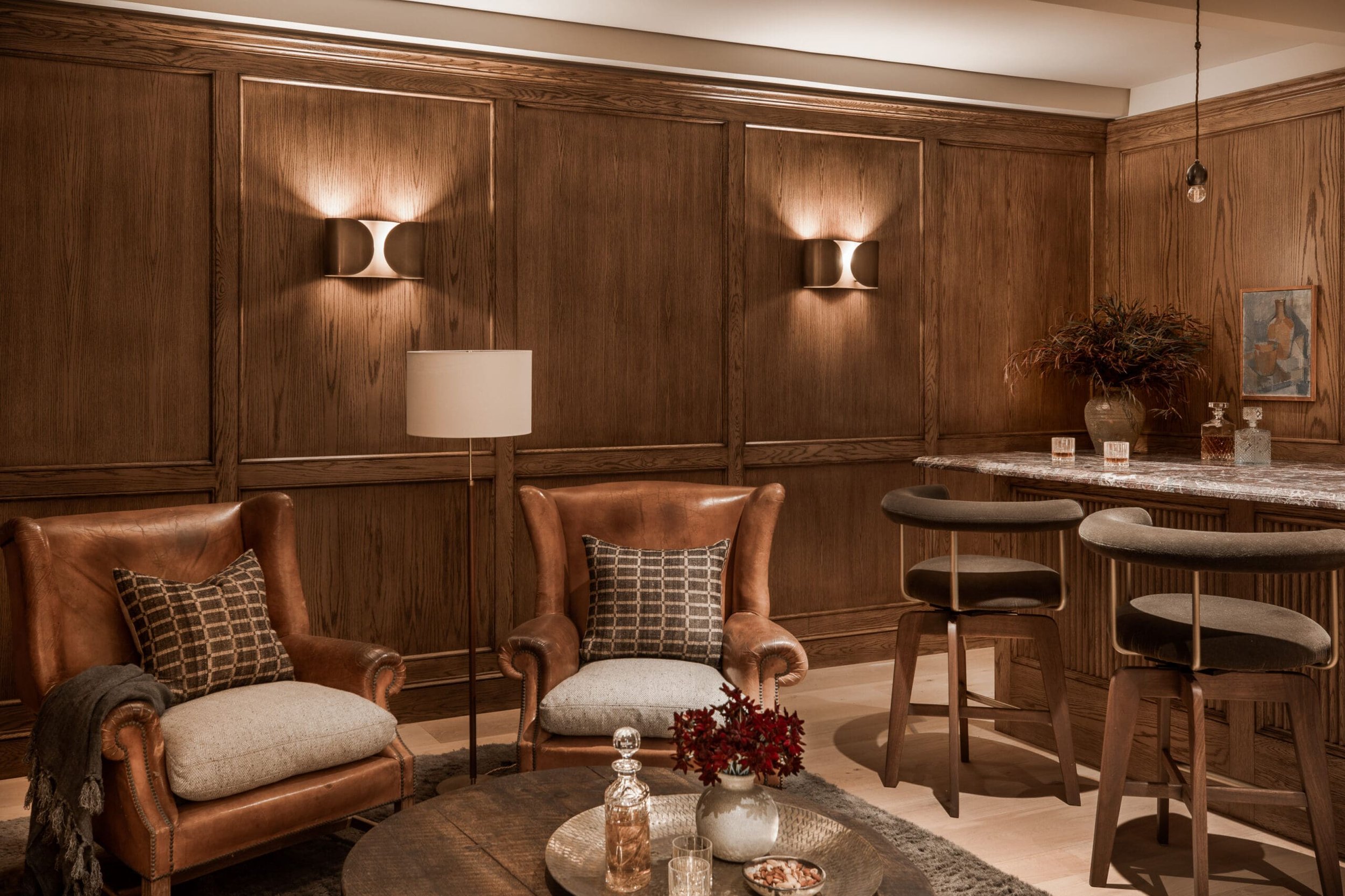 Living room with leather armchairs, a floor lamp, wood-paneled walls, a bar with stools, a floral arrangement, and artwork.