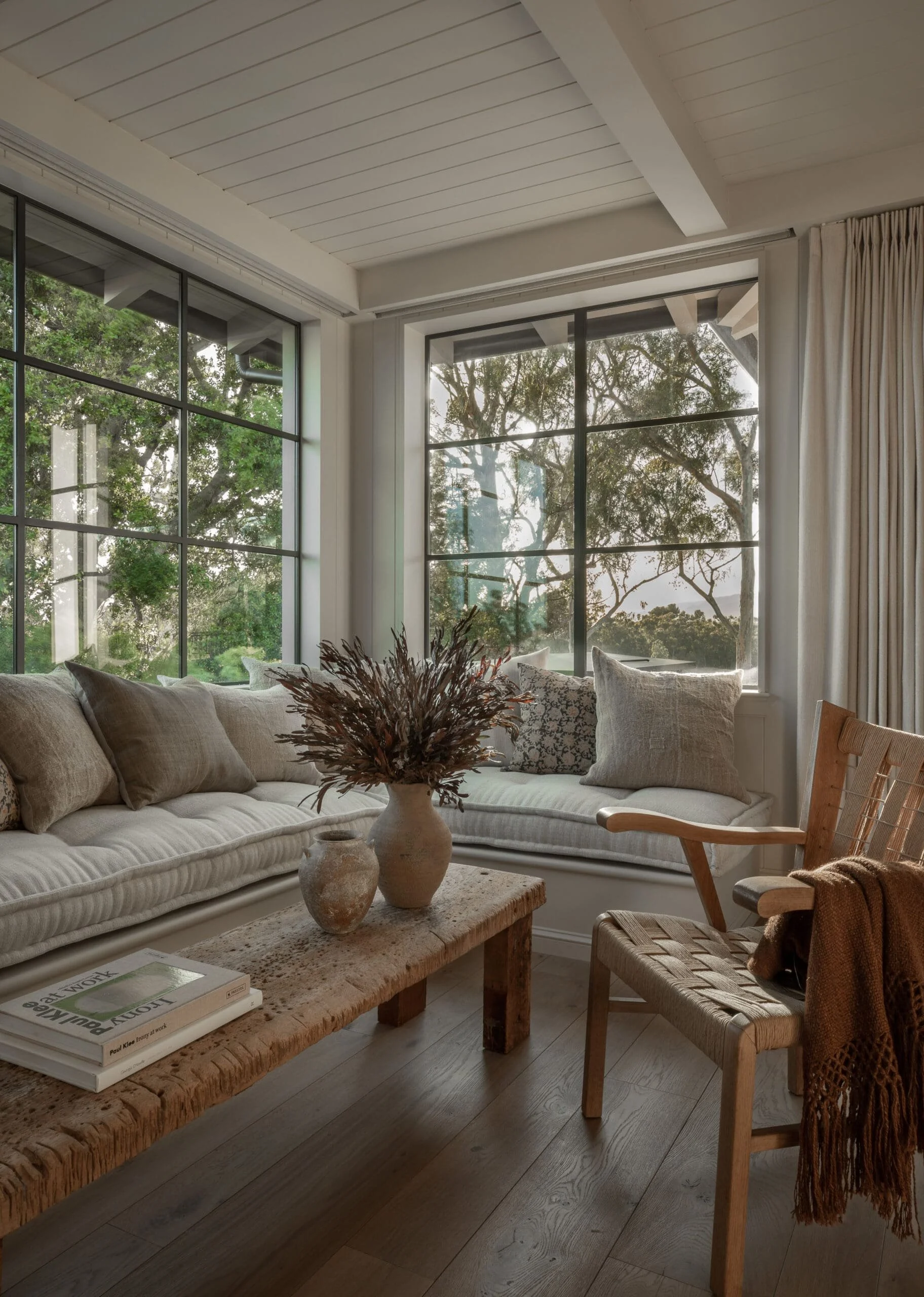 A cozy living room with large windows showing green trees outside. Contains a beige cushioned sofa with multiple pillows, a wooden coffee table with vases and a book, and a wooden chair with a brown throw blanket.