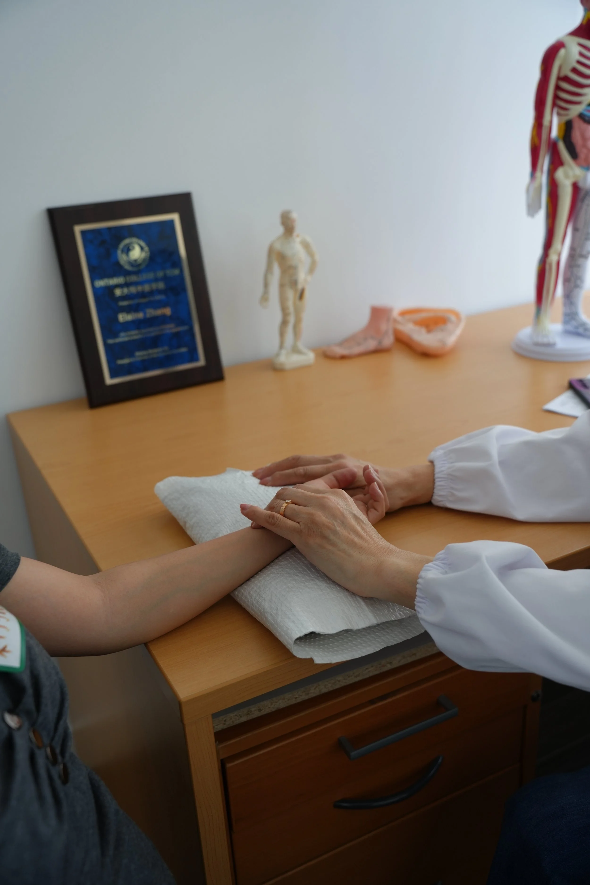 A woman receiving acupuncture treatment from a healthcare professional in a treatment room with a wooden table, lamp, and traditional Chinese medicine chart.