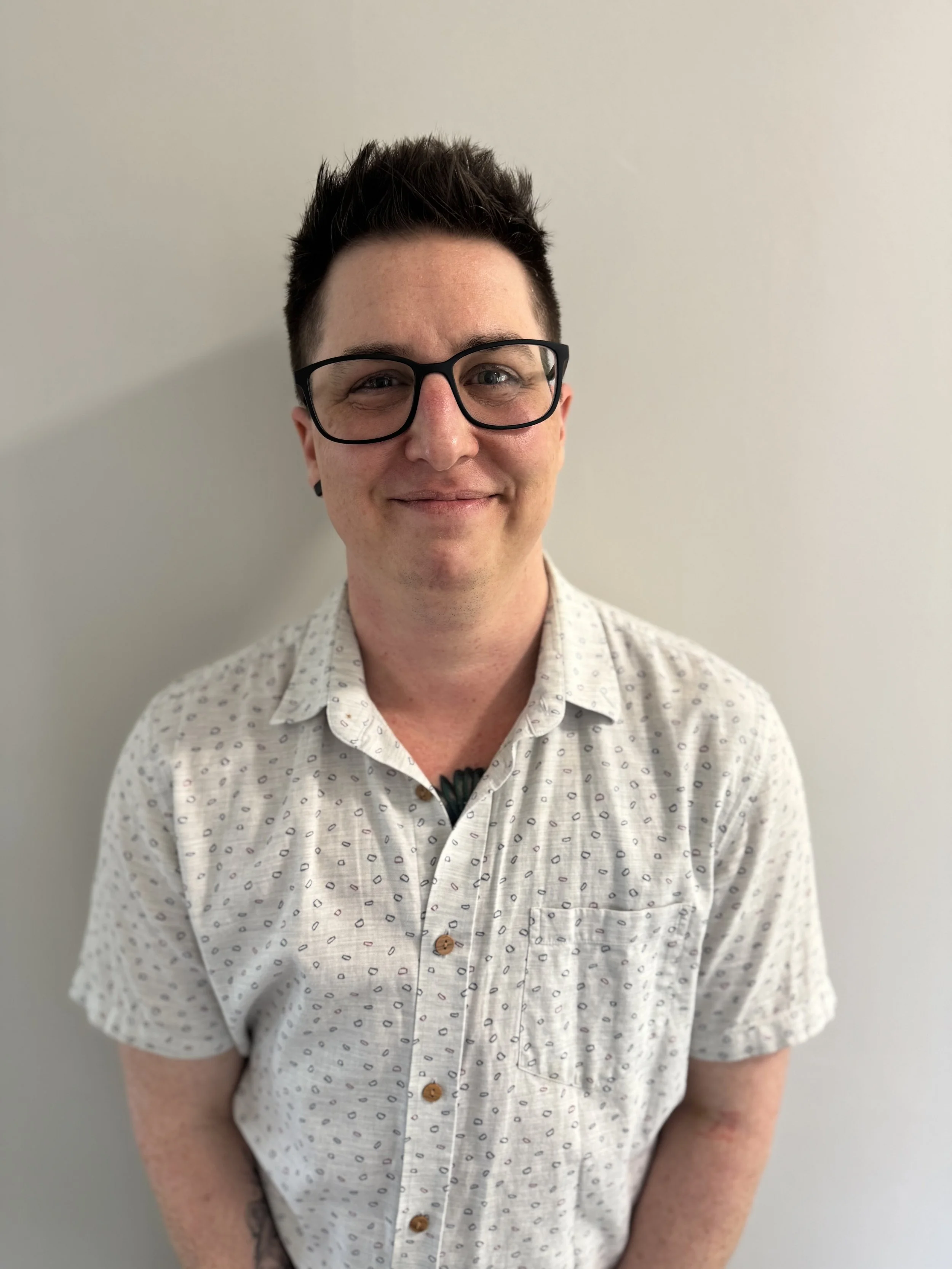 The image is of Dr Lucas Edwards standing facing the camera with a white background. They have short dark brown hair, black-framed glasses and a white short-sleeved collared shirt with small designs on it.