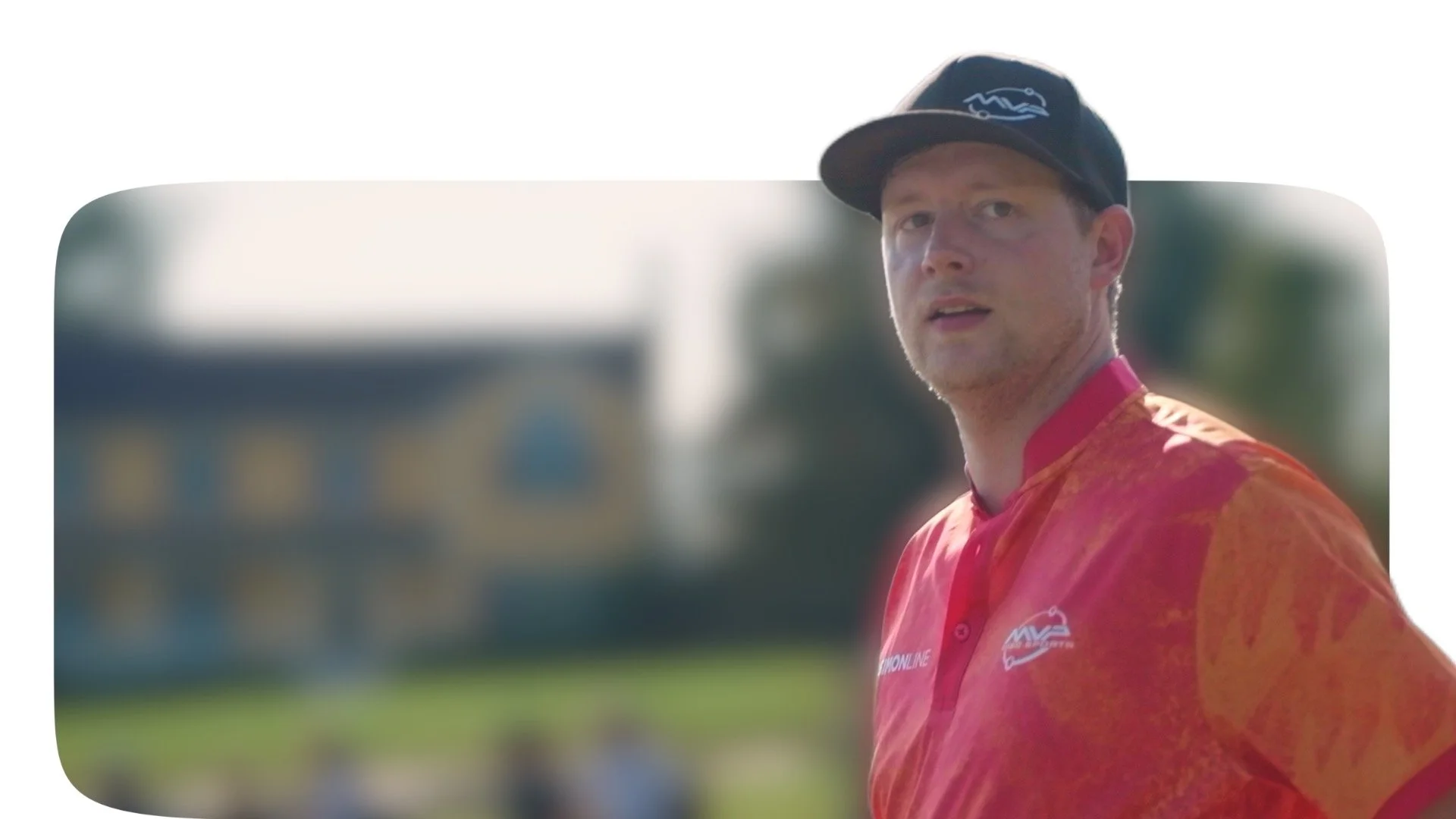 A man wearing a black cap and red sports jersey with orange and yellow patterns, standing outdoors with a blurred building and greenery in the background.