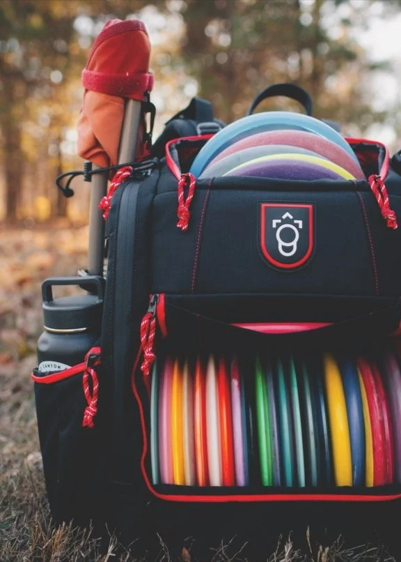 Backpack filled with colorful frisbees and a golf club, set outdoors on the ground with trees in the background.