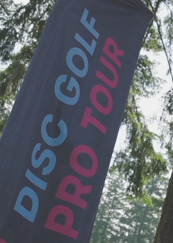 A flag with the words "DISCO GOLF PRO TOUR" printed on it, set outdoors among trees.