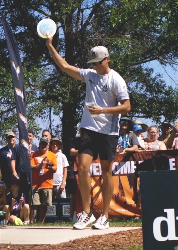 A young man wearing a white T-shirt, black shorts, and a cap is seen throwing a frisbee during an outdoor event, with a crowd of spectators watching.